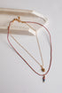 A delicate layered necklace set featuring a gold chain with a small gold spiral pendant and a longer red-and-gold beaded chain with a small multicolor fish charm, displayed on a light gray surface next to a white ceramic tray.