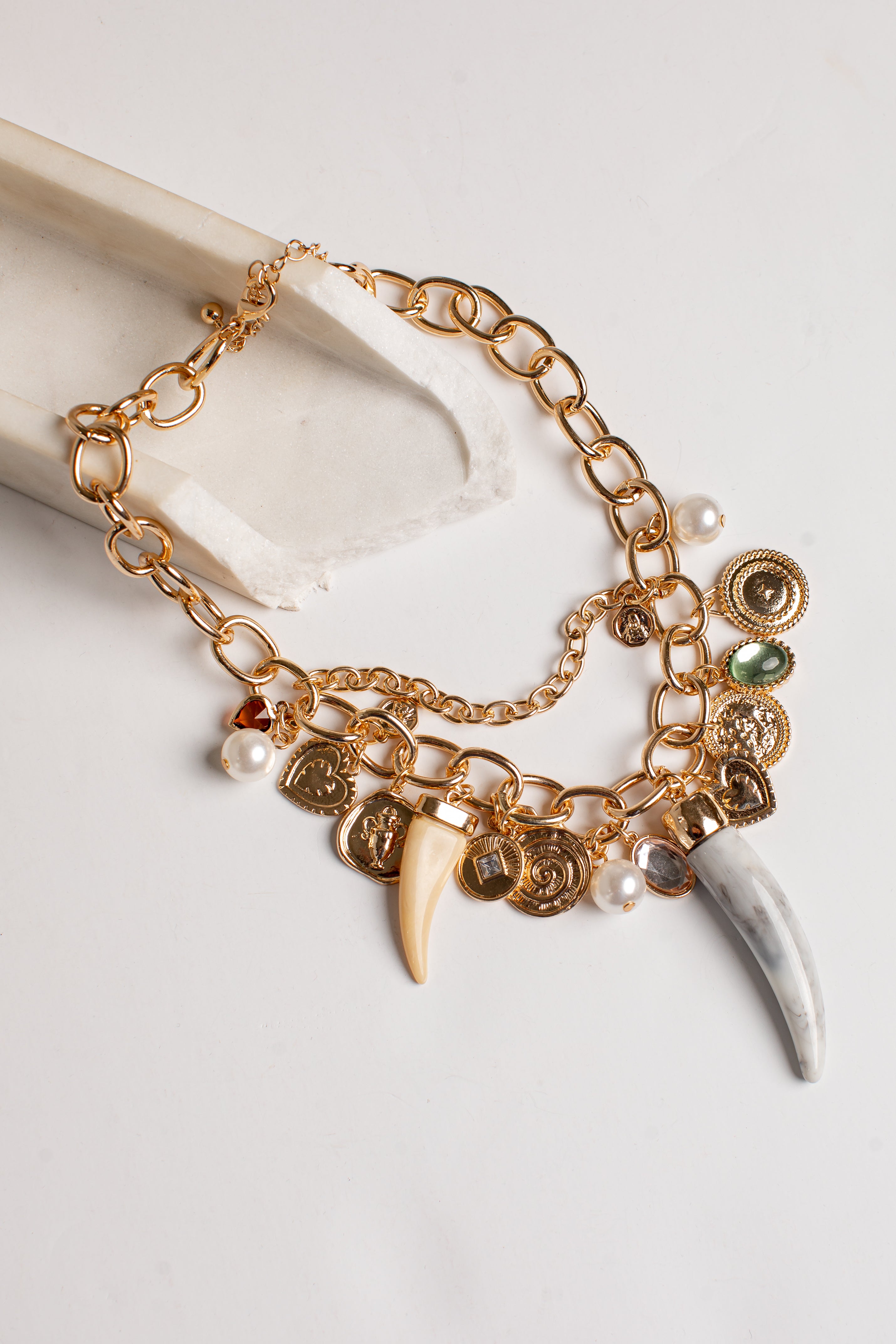 A gold-toned statement necklace featuring chunky chain links, small coin charms, pearl beads, a cream-colored tooth charm, and a larger white curved bone or horn charm, displayed on a light neutral surface.