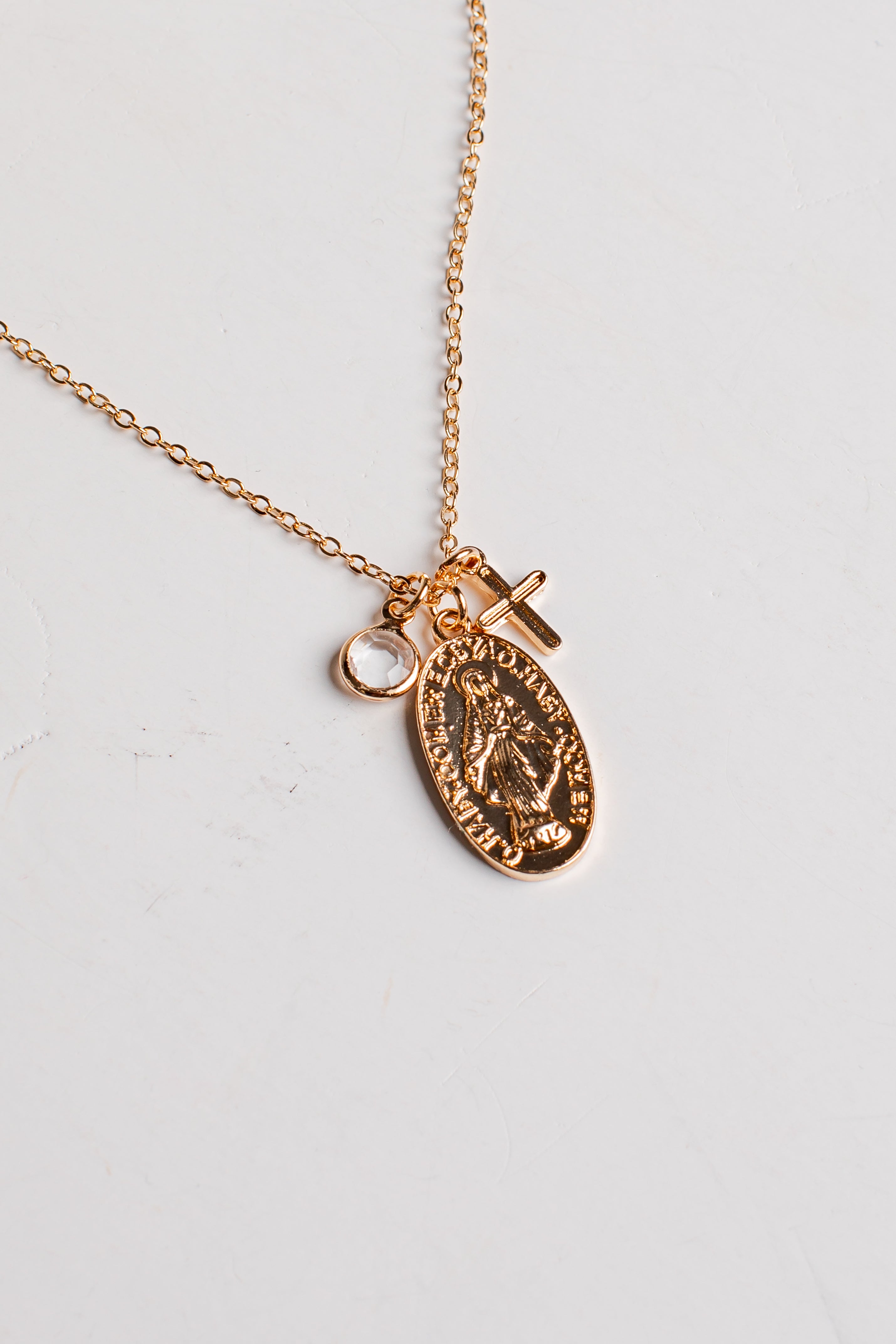 A rose gold chain necklace featuring an oval religious medallion pendant and a small cross charm, displayed on a light off-white background.