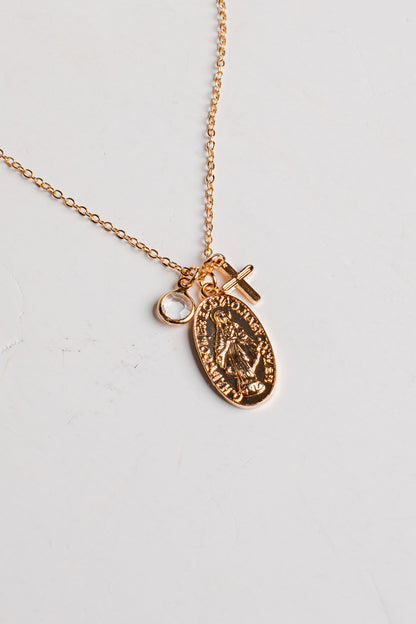 A rose gold chain necklace featuring an oval religious medallion pendant and a small cross charm, displayed on a light off-white background.