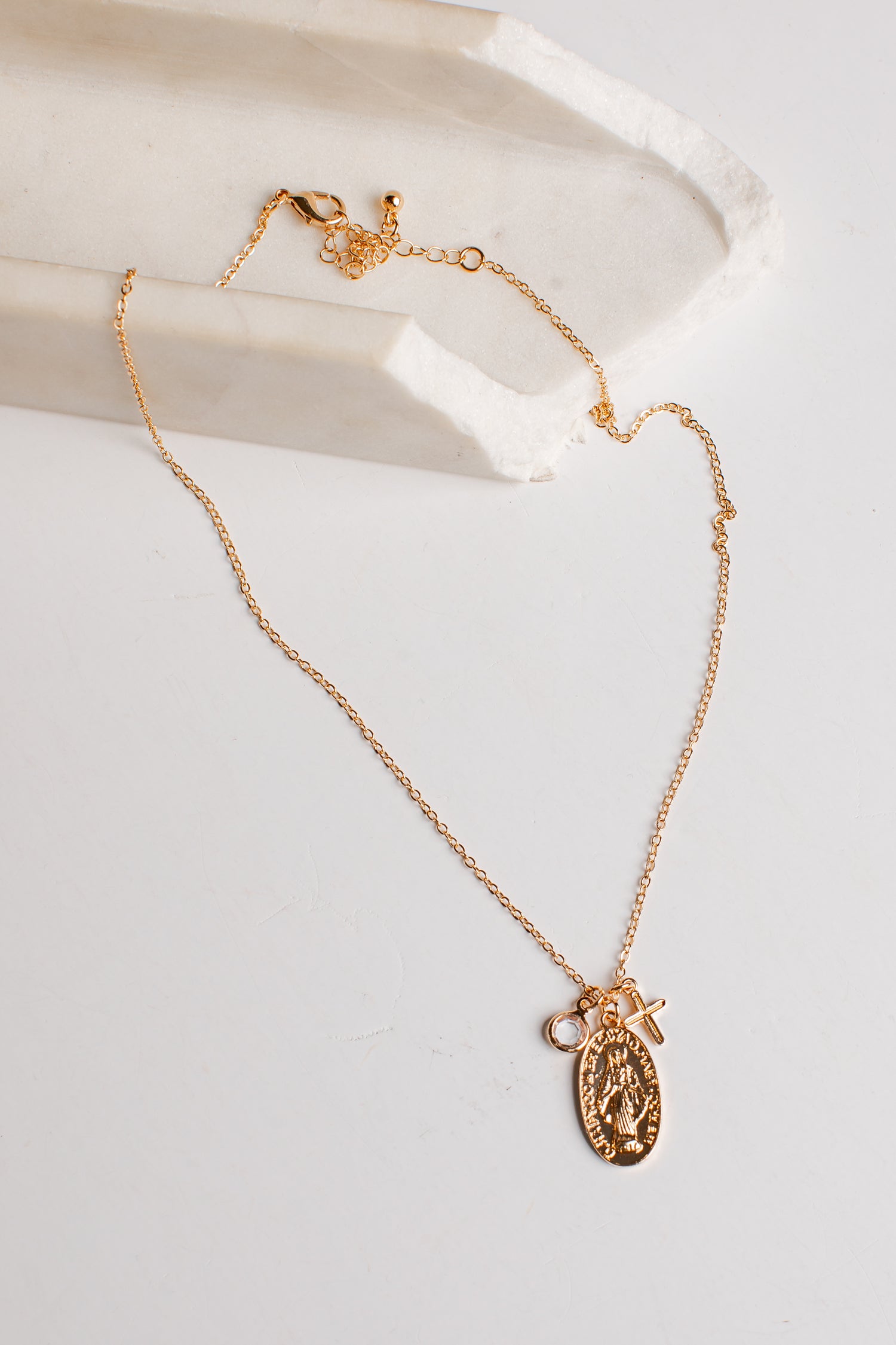A delicate gold chain necklace with a Saint Benedict medallion and a small cross charm, displayed on a white marble tray against a clean white background.