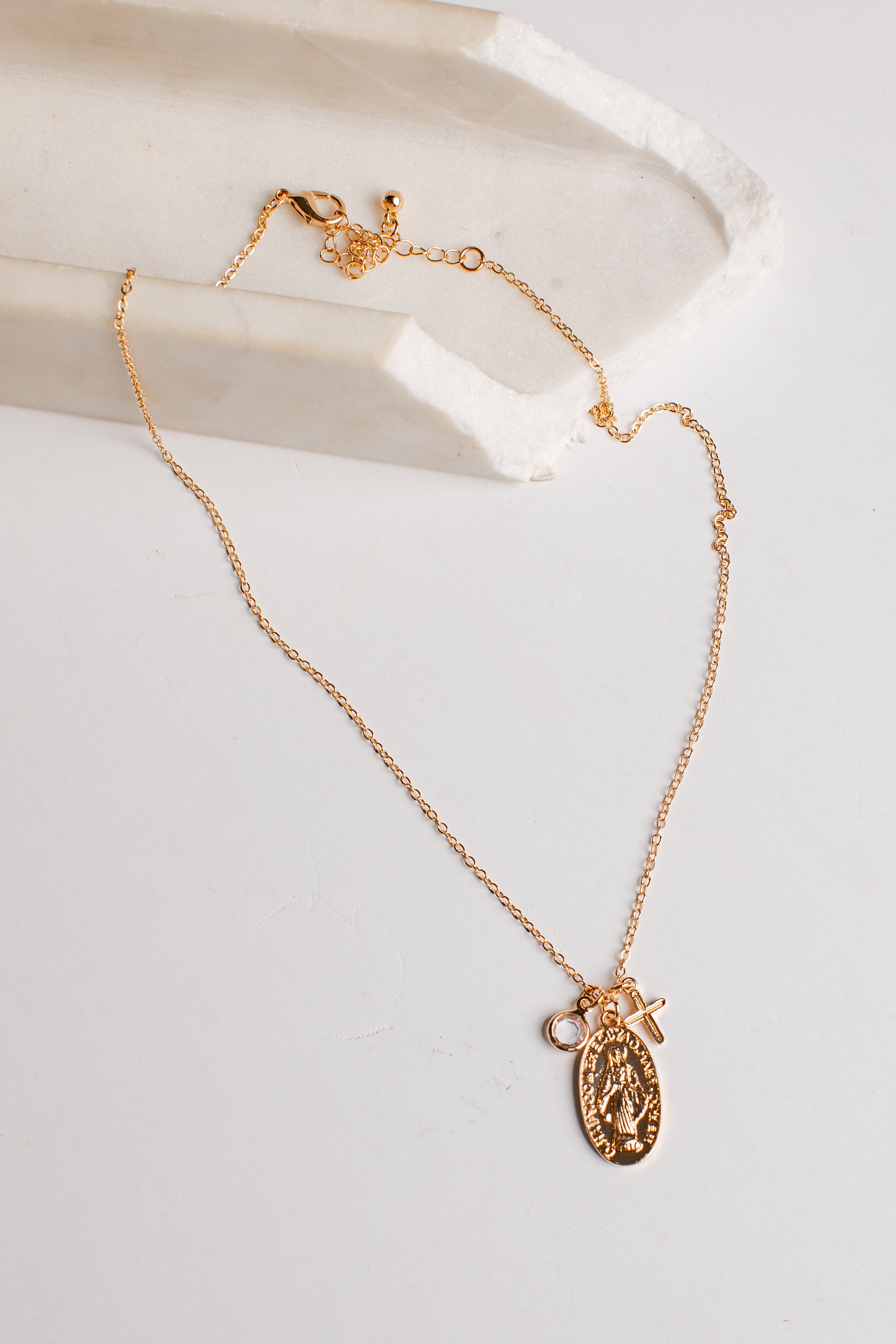 A delicate gold chain necklace with a Saint Benedict medallion and a small cross charm, displayed on a white marble tray against a clean white background.