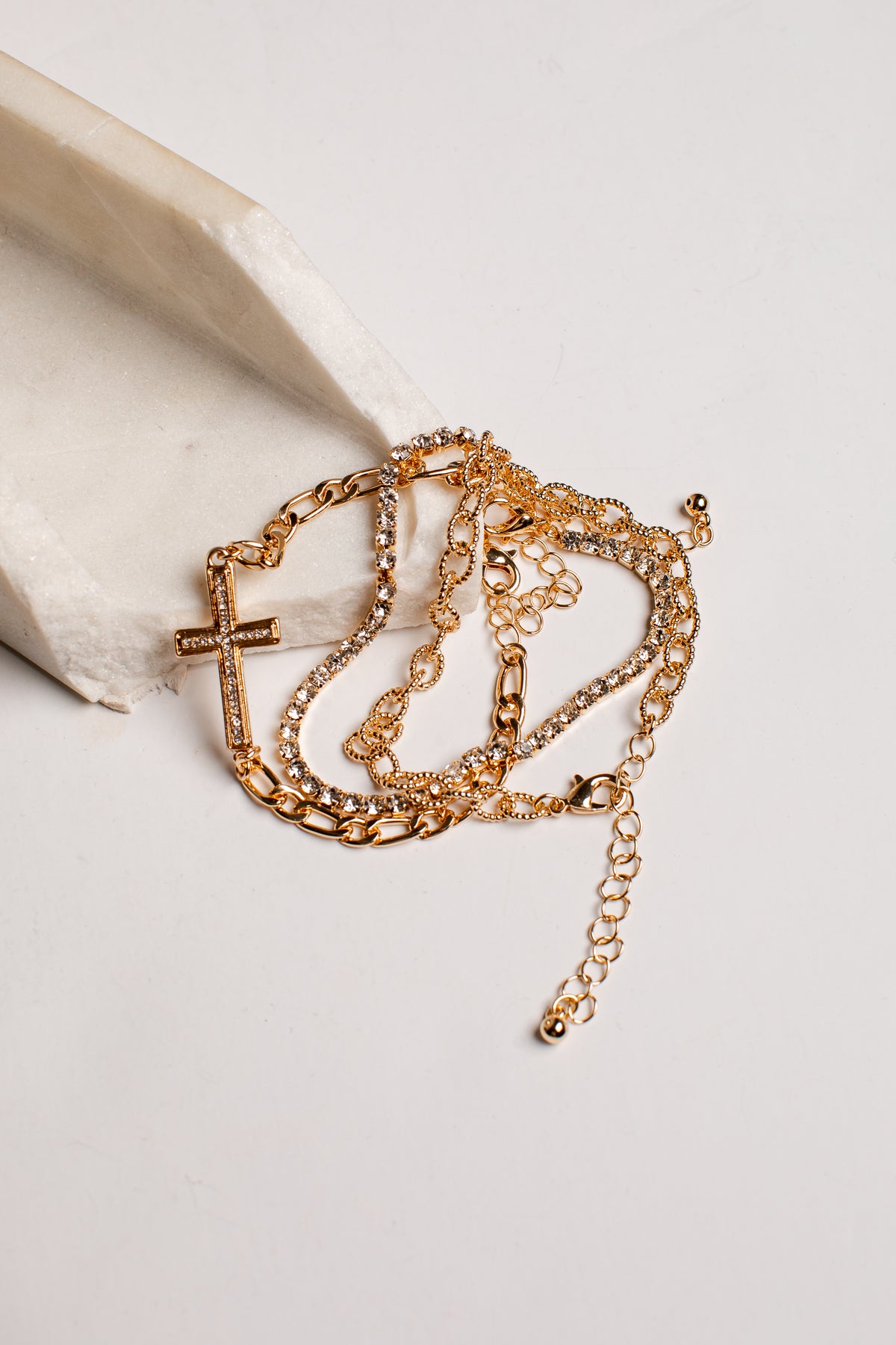 A layered gold-tone chain bracelet set with a cross pendant and textured beaded links, displayed on a light cream stone prop against a white background.