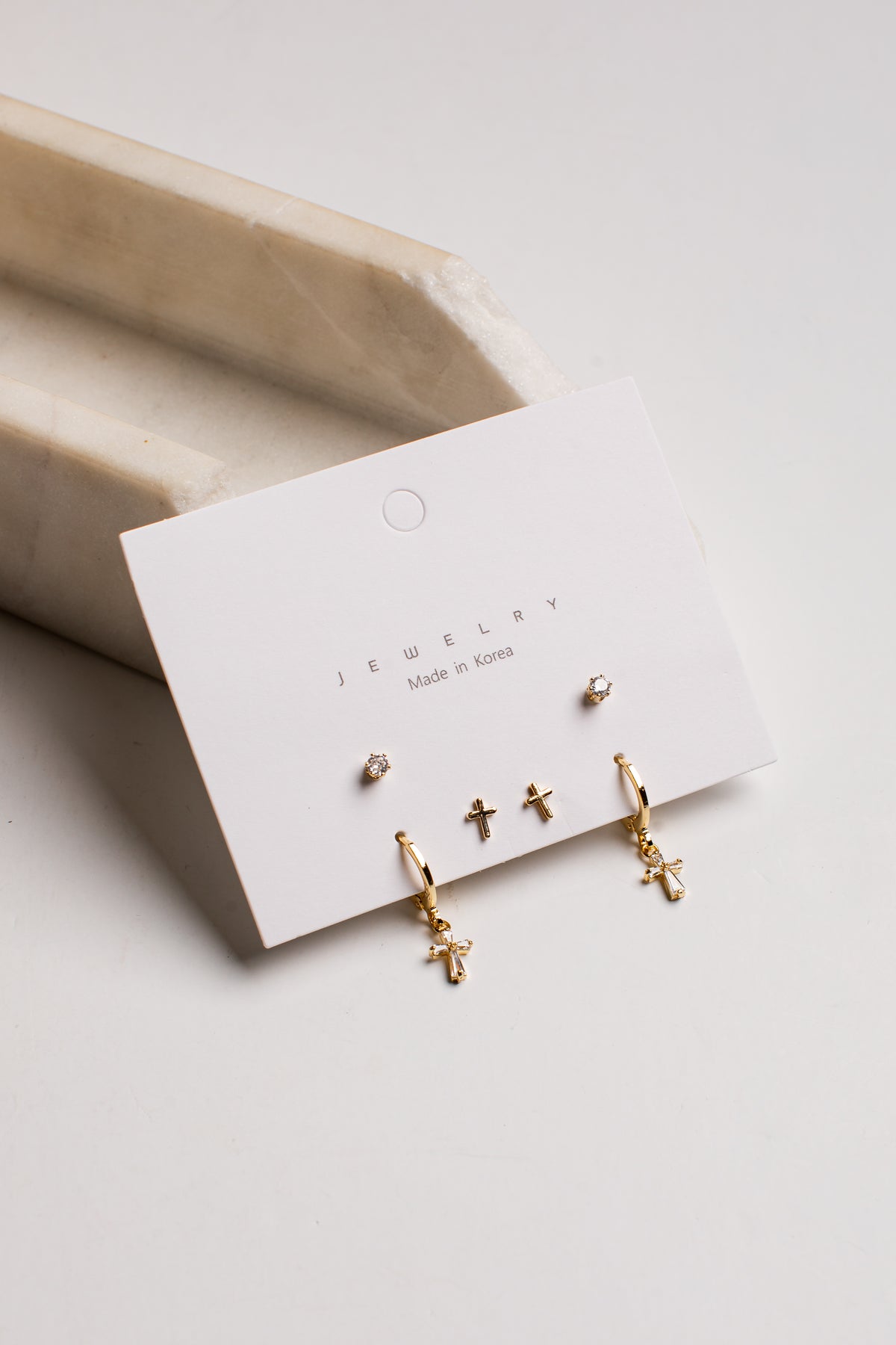A set of gold-toned stud and hoop earrings displayed on a white jewelry card, photographed on a light neutral surface with a beige tray in the background.