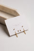 A set of gold-toned stud and hoop earrings displayed on a white jewelry card, photographed on a light neutral surface with a beige tray in the background.