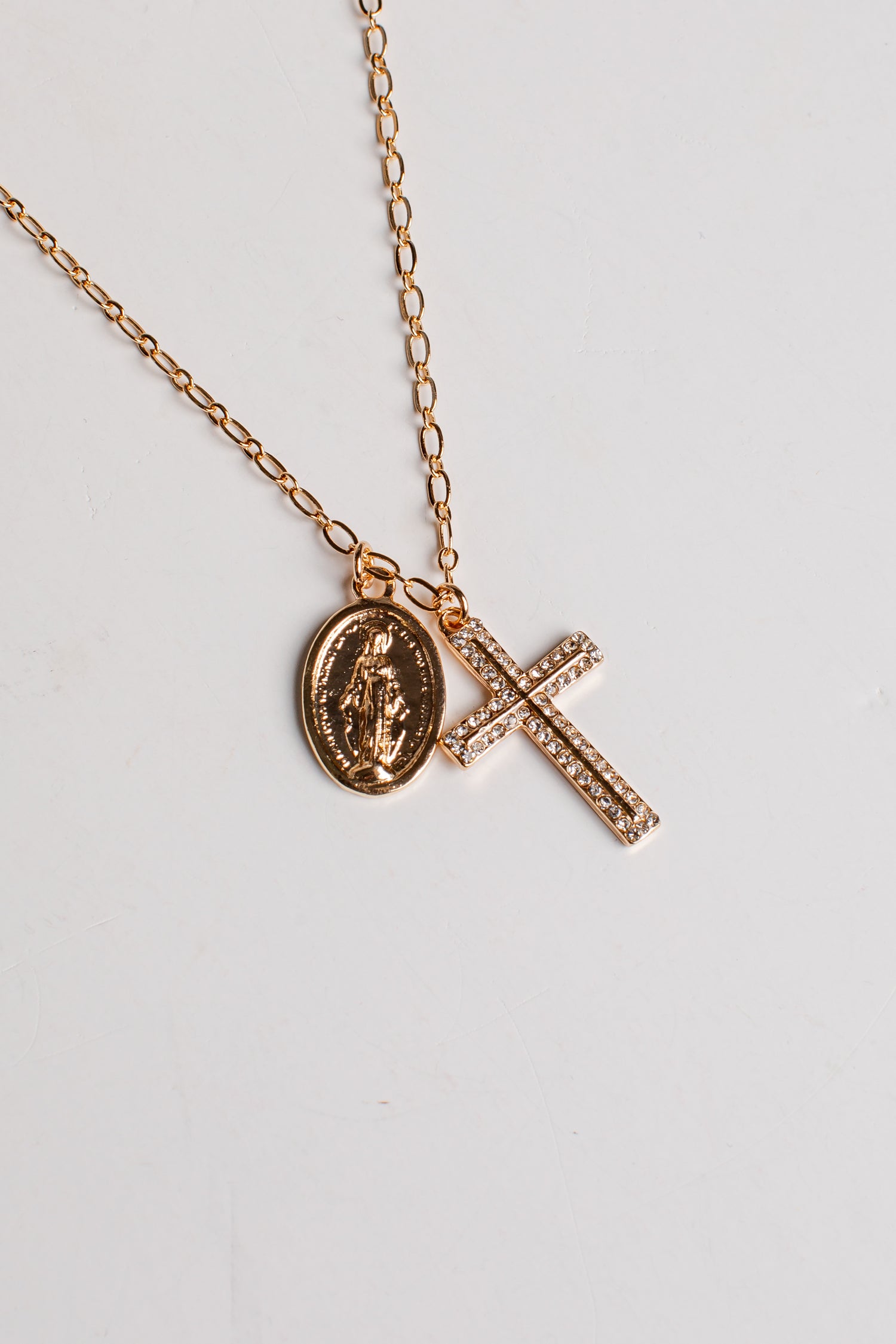 A rose gold chain necklace featuring two pendants: an oval Miraculous Medal pendant and a rectangular cross paved with small clear stones, displayed on a light gray background.