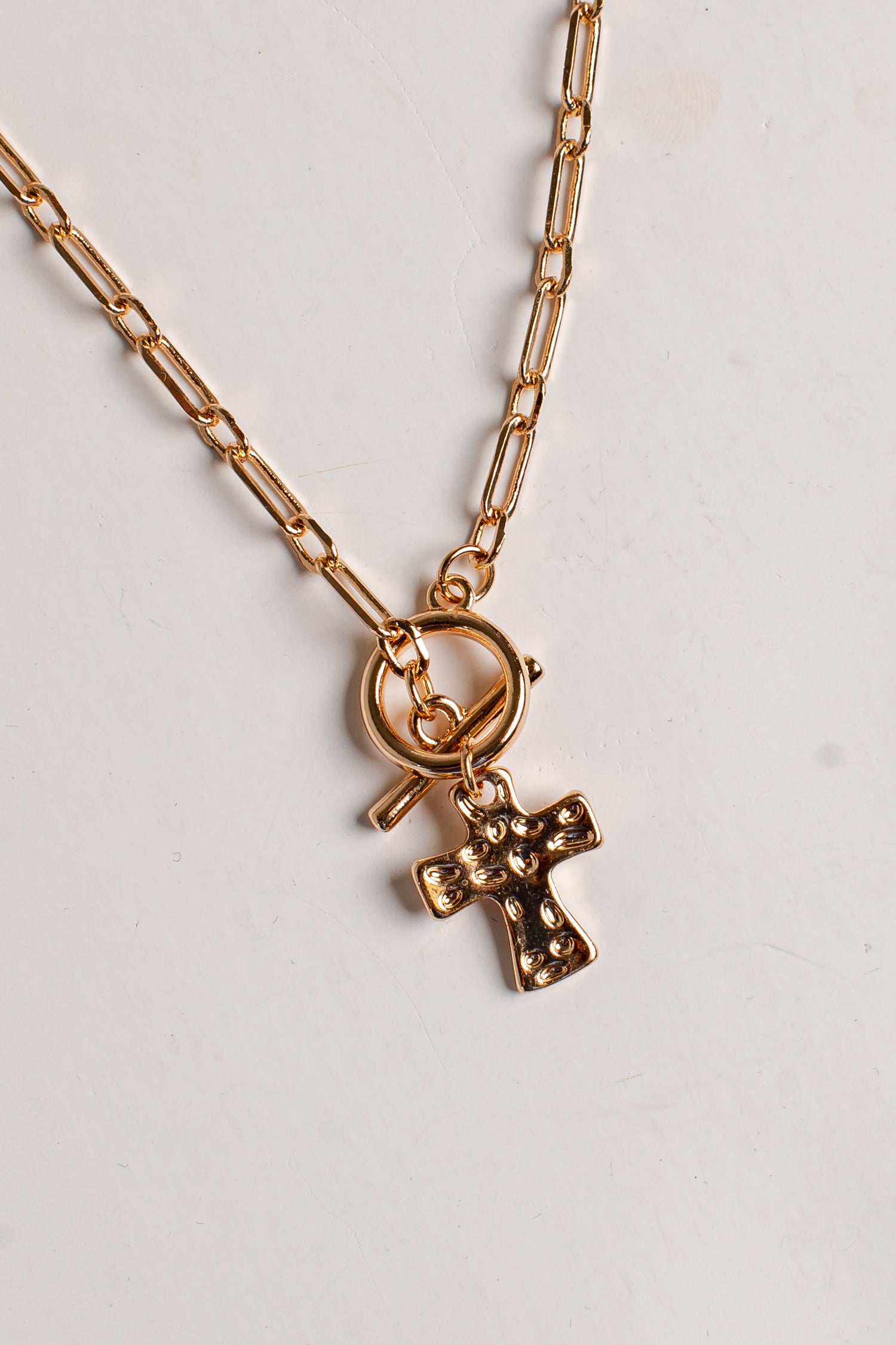 A gold-toned paperclip chain necklace with a small cross pendant featuring tiny clear rhinestones, displayed on a light off-white background.