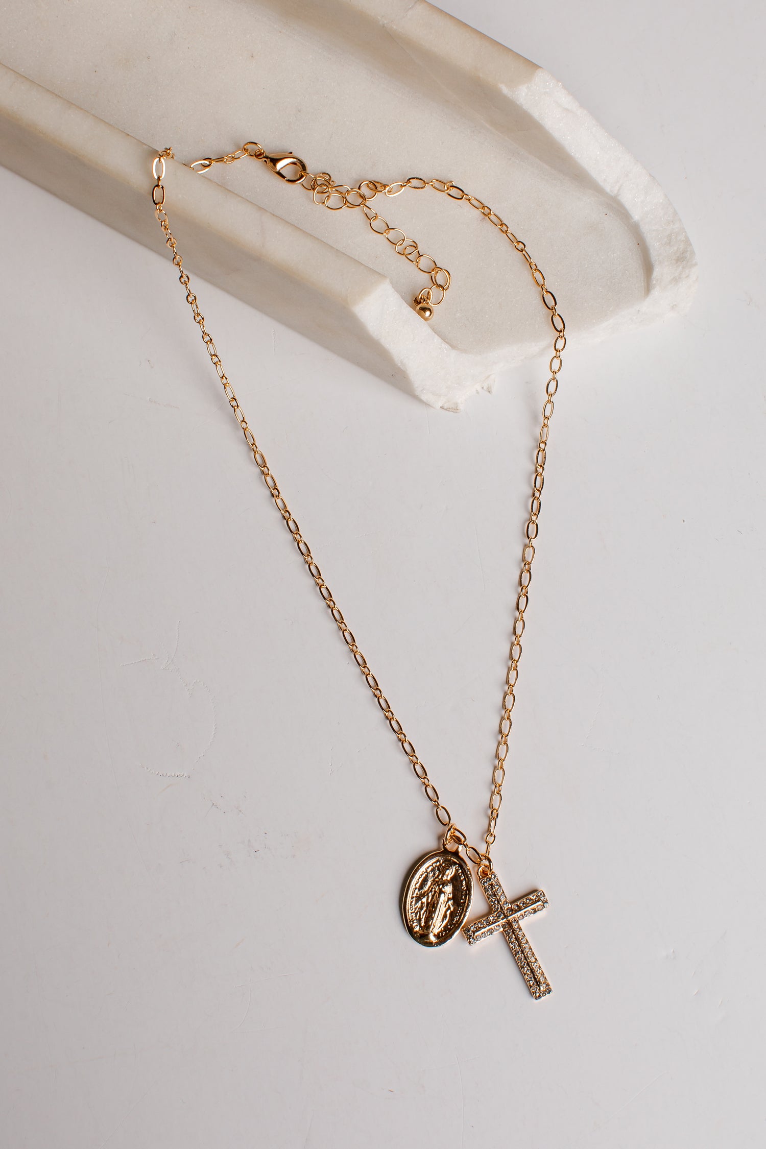A delicate gold chain necklace featuring a small oval religious medallion and a textured cross pendant, displayed on a white marble tray against a light gray background.