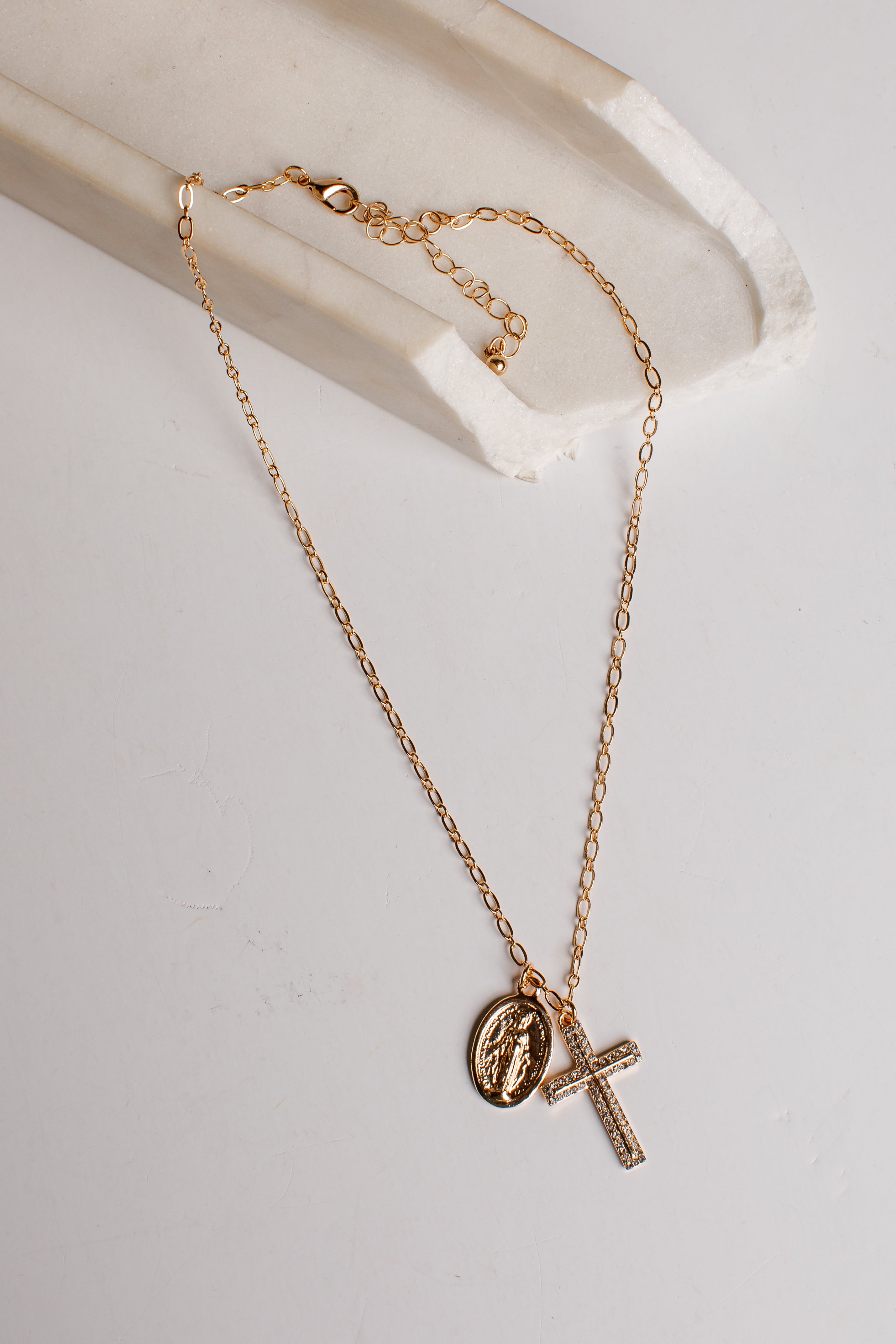 A delicate gold chain necklace featuring a small oval religious medallion and a textured cross pendant, displayed on a white marble tray against a light gray background.
