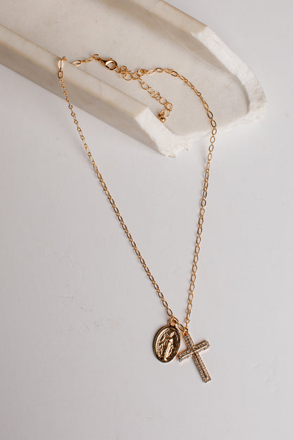 A delicate gold chain necklace featuring a small oval religious medallion and a textured cross pendant, displayed on a white marble tray against a light gray background.