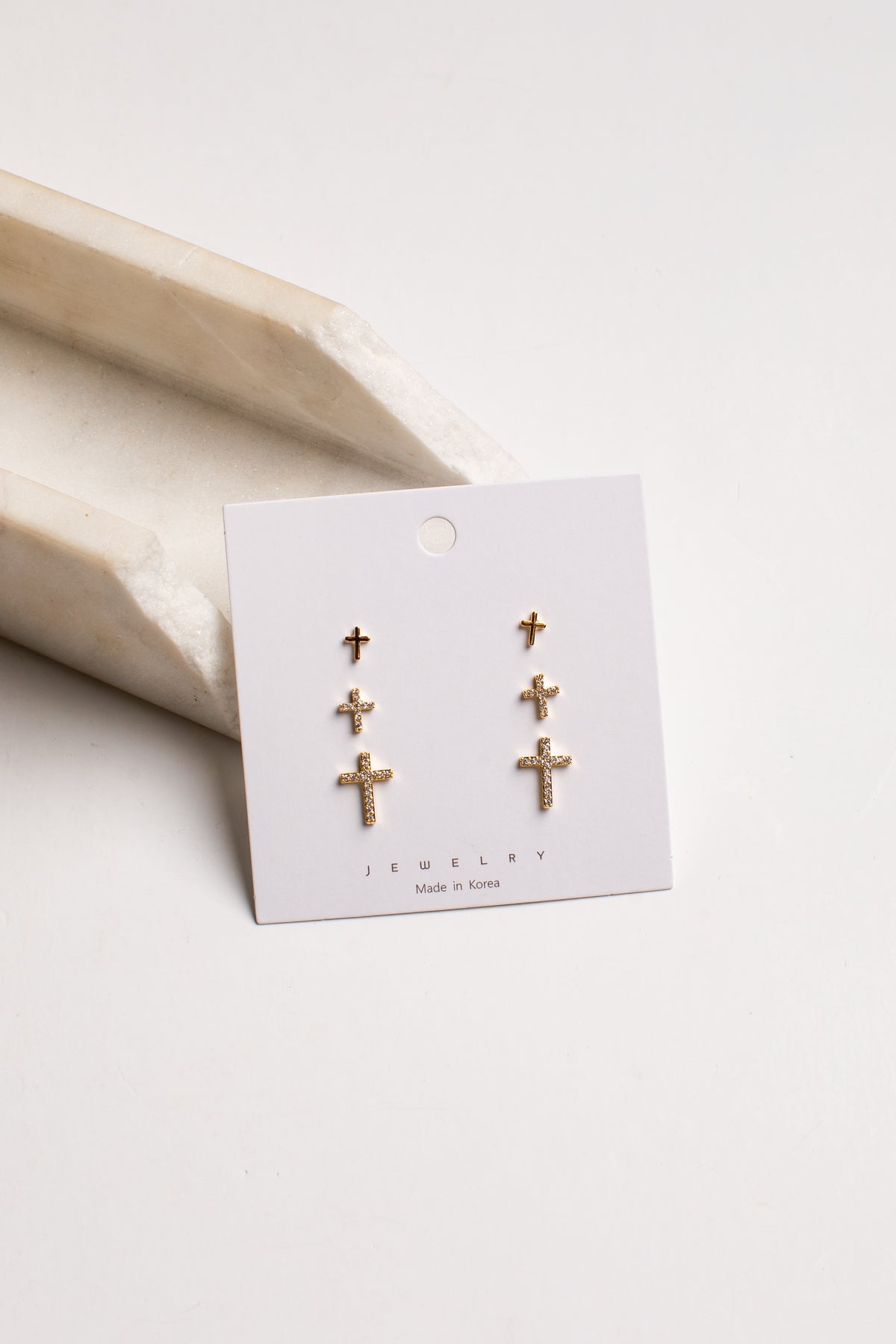 A pair of gold cross stud earrings displayed on a white jewelry card, resting on a light beige stone prop against a clean white background.