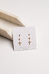 A pair of gold cross stud earrings displayed on a white jewelry card, resting on a light beige stone prop against a clean white background.