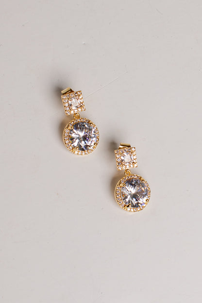 A pair of gold drop earrings with clear round-cut stones, featuring a small pavé-set square top and a larger round halo-set drop, displayed on a light gray background.