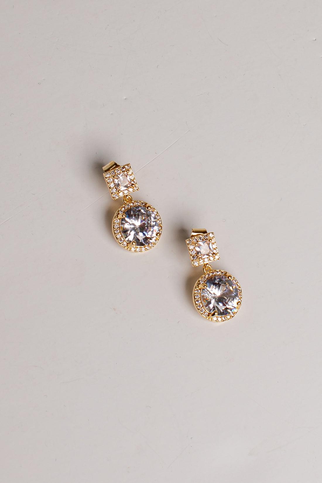 A pair of gold drop earrings with large clear round gemstones and smaller clear stones above them, photographed on a plain light gray background.
