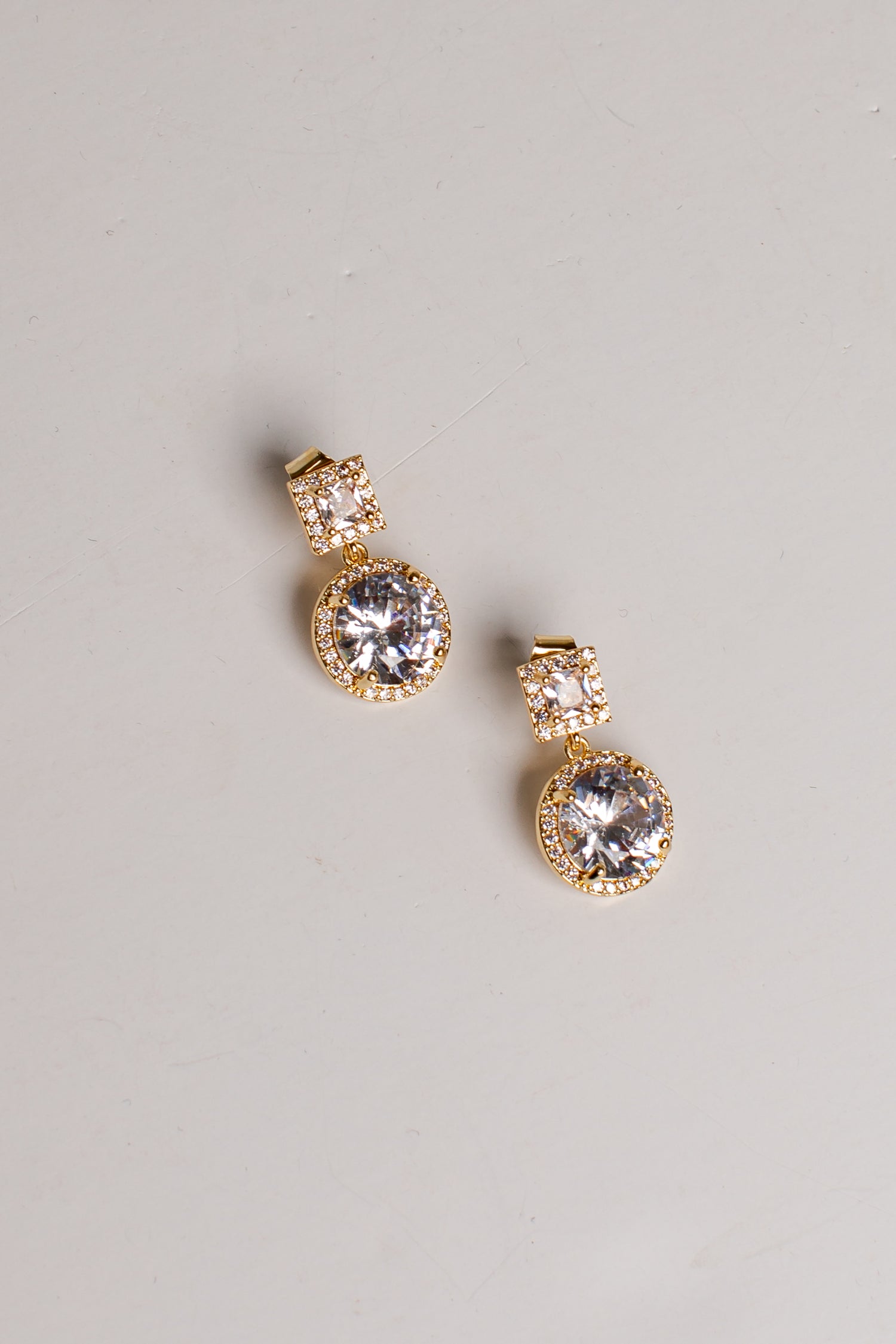 A pair of gold drop earrings with large clear round gemstones and smaller clear stones above them, photographed on a plain light gray background.