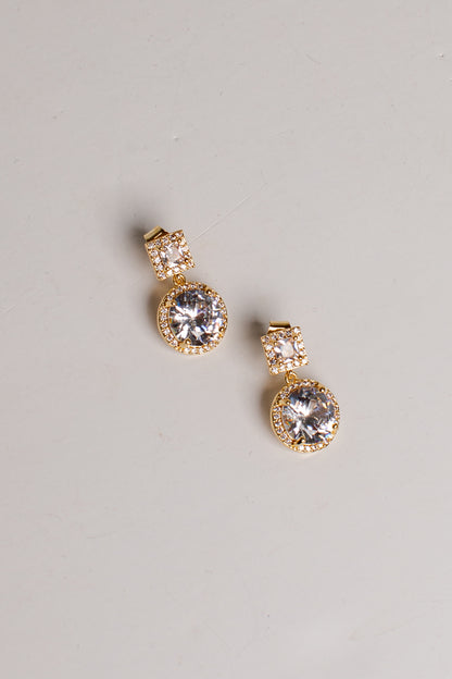 A pair of gold drop earrings with large clear round gemstones and smaller clear stones above them, photographed on a plain light gray background.