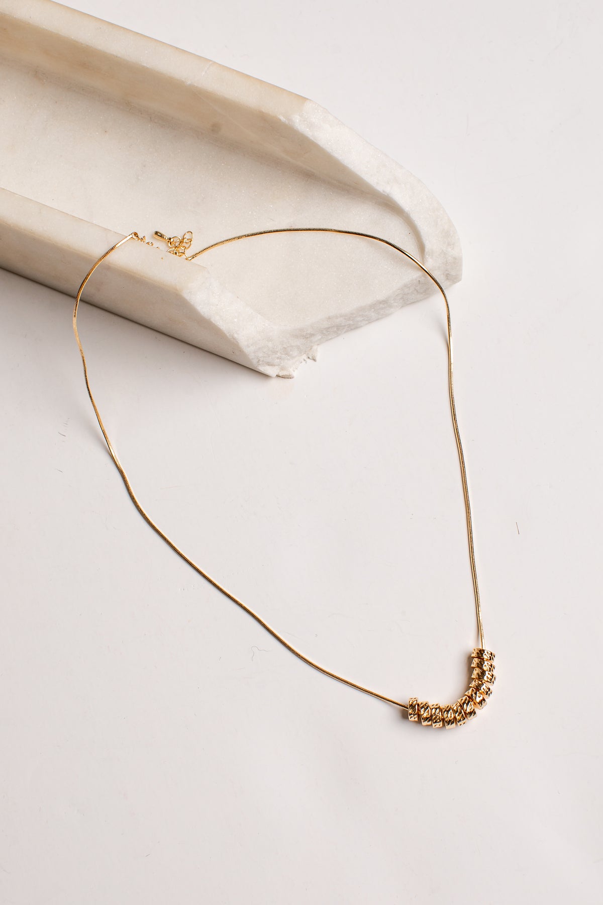 A delicate gold chain necklace with a small textured gold pendant, displayed on a light cream-colored stone riser against a clean white background.