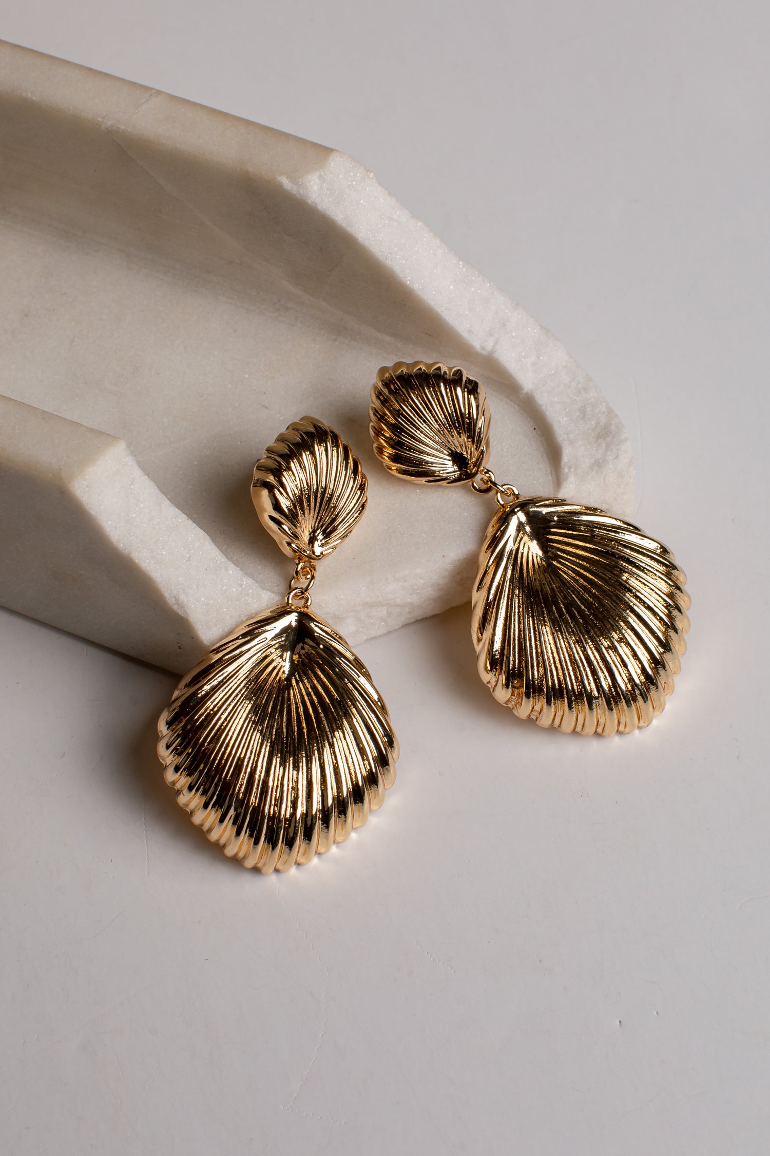A pair of gold-toned drop earrings with textured, ribbed shell-like dangles, displayed on a light gray surface next to a white stone prop.