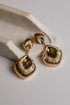 A pair of gold-toned drop earrings with textured, ribbed shell-like dangles, displayed on a light gray surface next to a white stone prop.