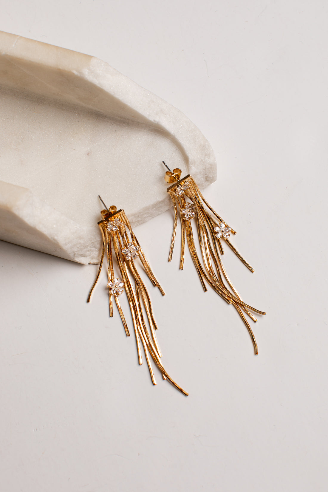 A pair of gold-toned dangling earrings with multiple thin fringe strands and small clear rhinestone clusters, displayed on a light neutral surface beside a curved cream stone prop.