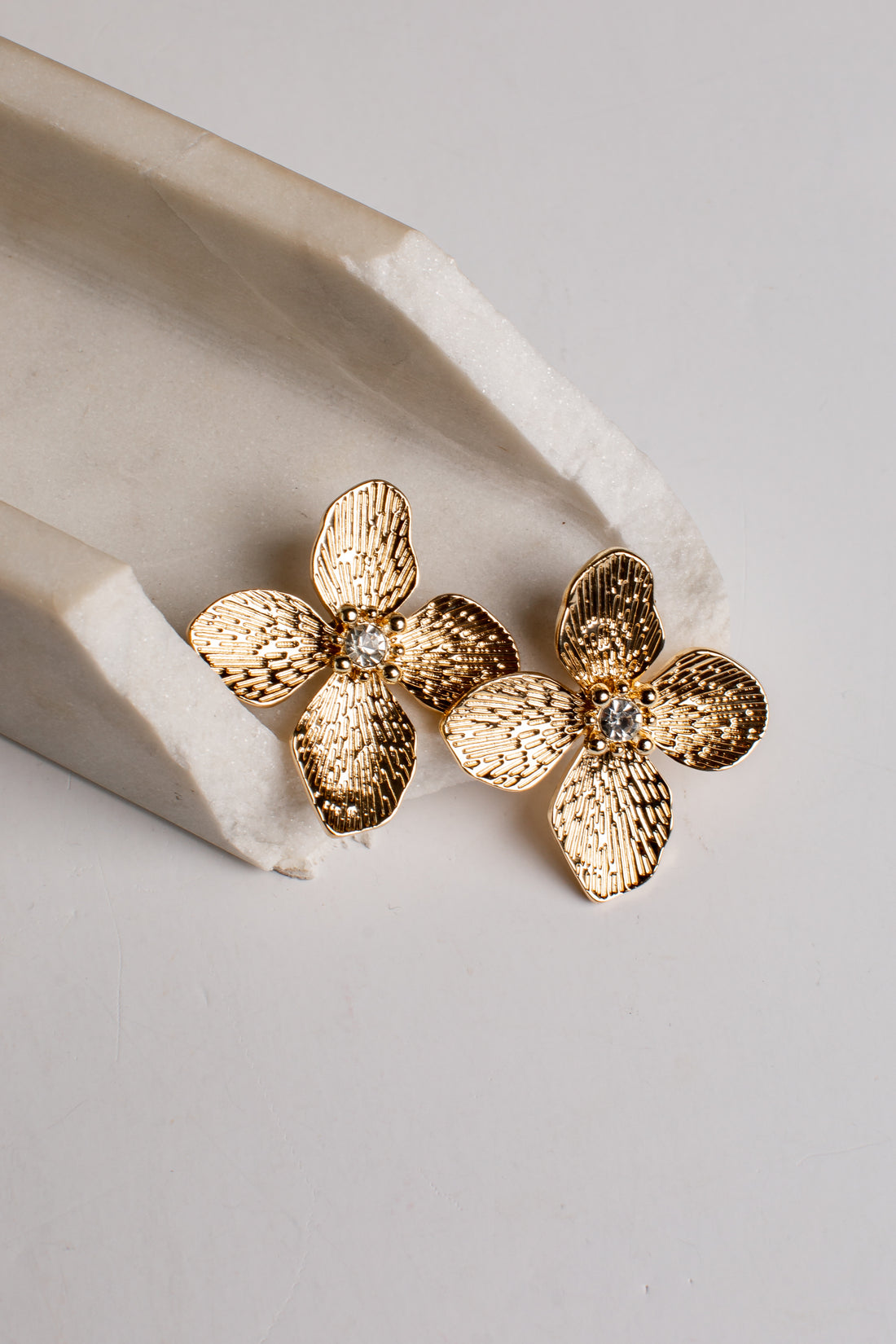 A pair of textured gold flower-shaped stud earrings with a small clear rhinestone in the center, resting on a light beige stone prop against a neutral light gray background.
