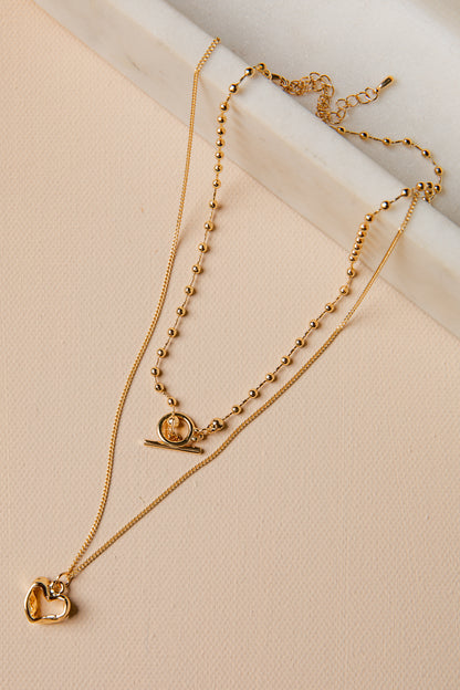 Two gold necklaces with heart and circular pendants on a beige surface