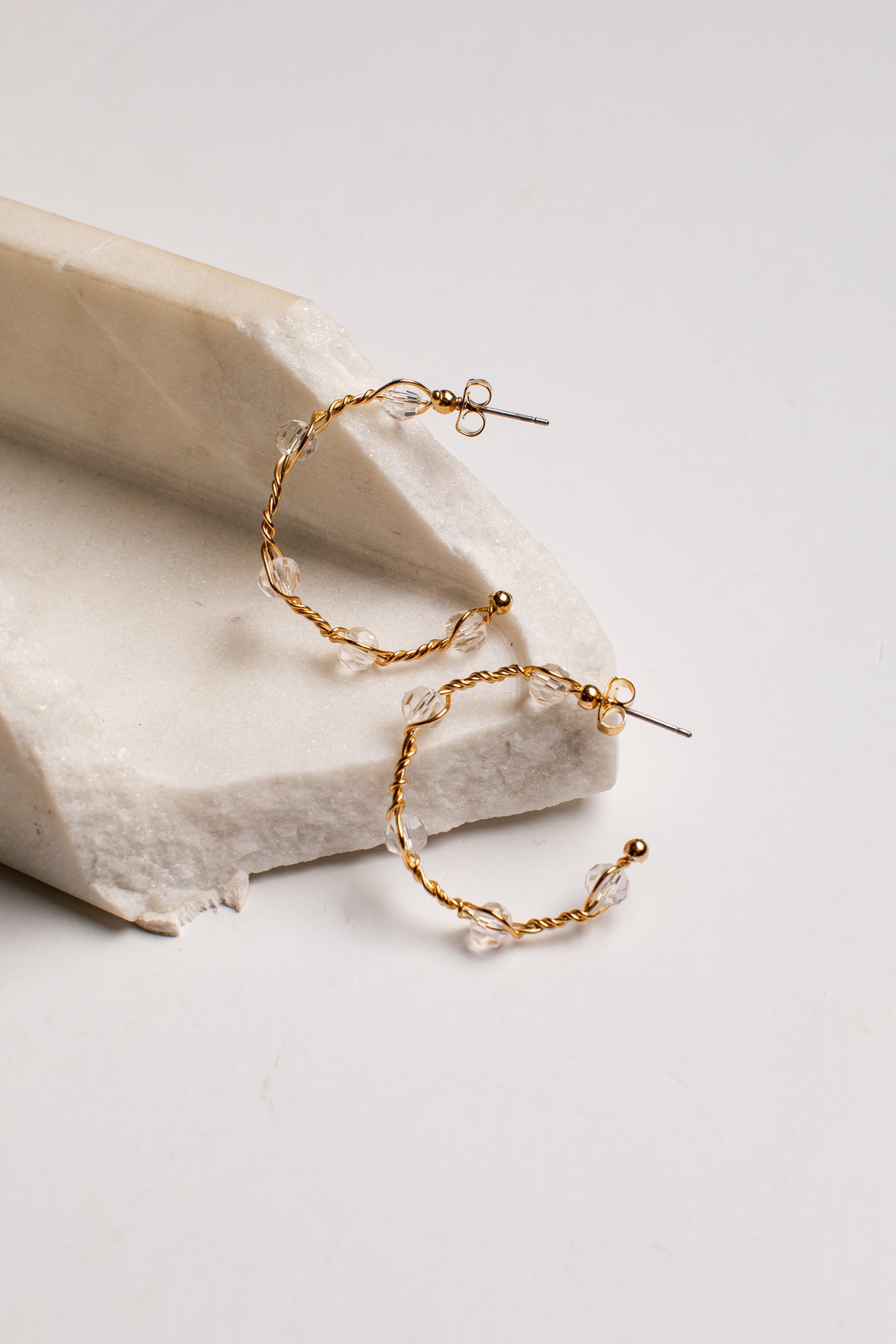 A pair of gold hoop earrings with clear faceted beads, displayed on a textured white stone riser against a clean white background.