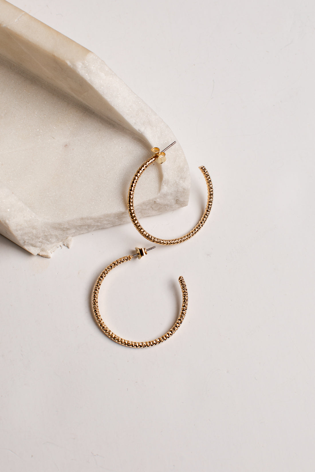 A pair of textured gold hoop earrings displayed on a light beige stone slab against a clean off-white background.