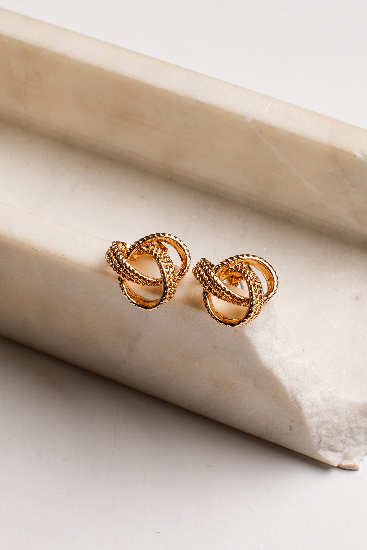 A pair of gold-toned knot stud earrings resting on a textured beige stone slab against a clean white background.