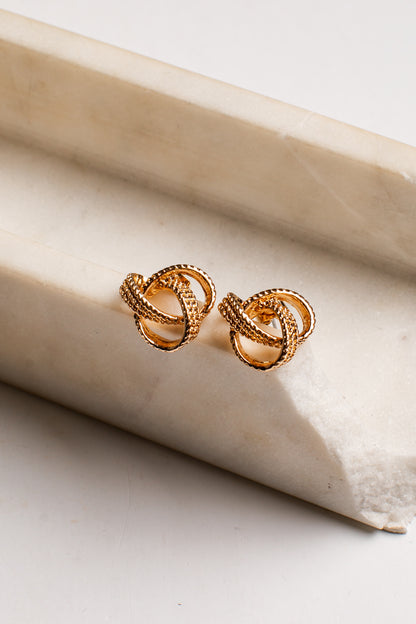 A pair of gold-toned knot stud earrings resting on a textured beige stone slab against a clean white background.