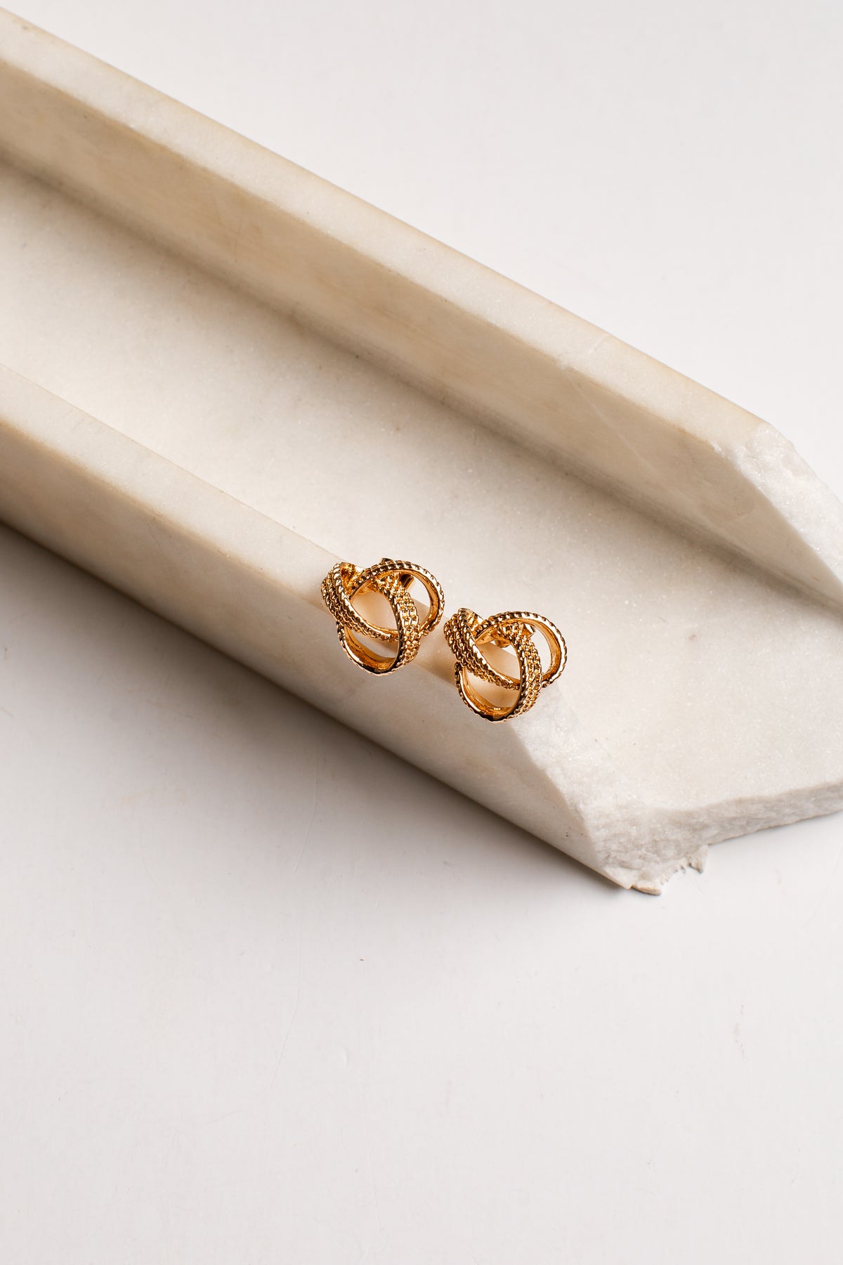 A pair of small gold knot stud earrings resting on a cream-colored stone tray against a white background.