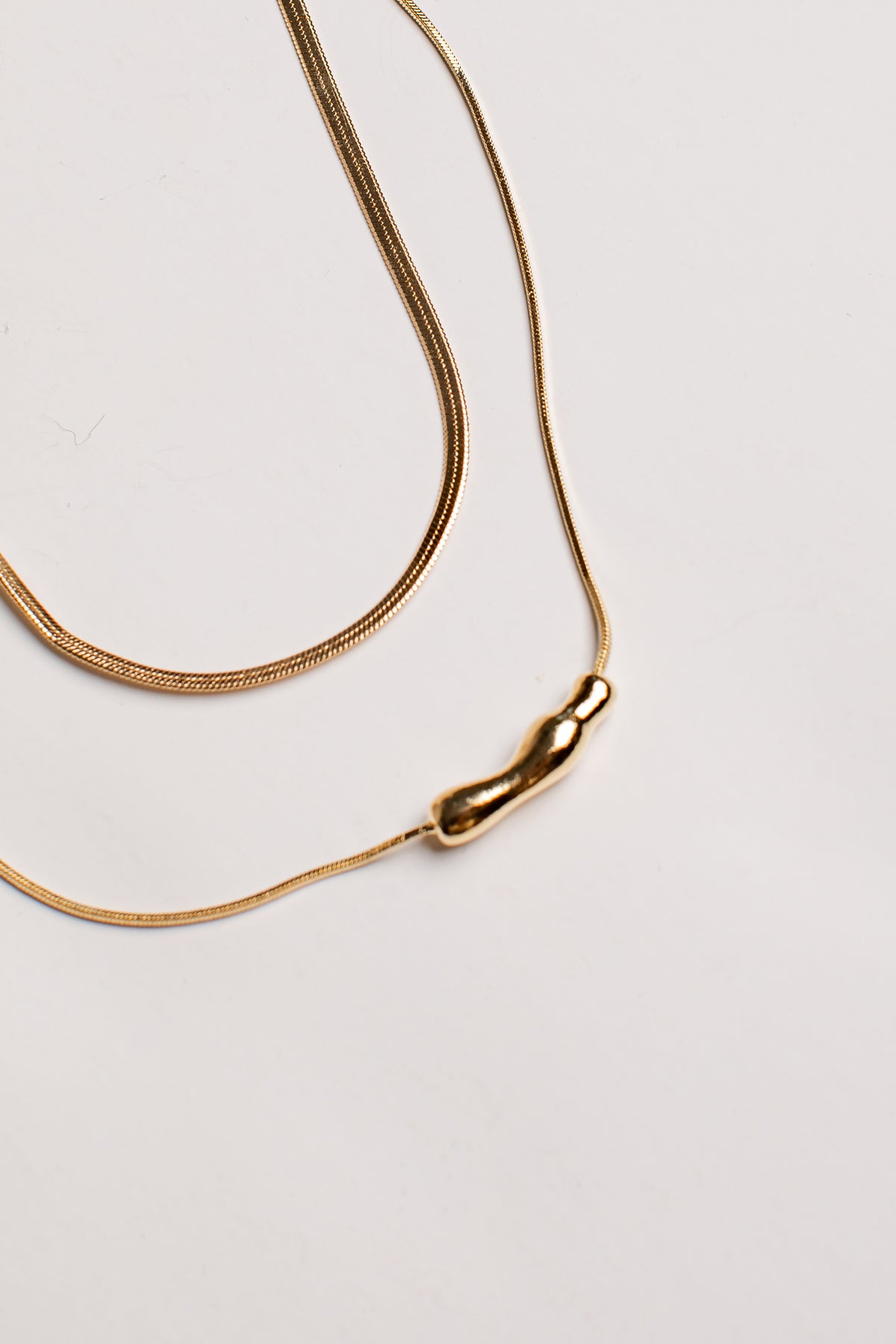 A gold snake chain necklace with a small, polished gold bead pendant, photographed on a clean, light off-white background.