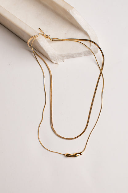 A delicate gold chain necklace with a small polished gold pendant, draped elegantly over a textured white stone block on a clean white background.