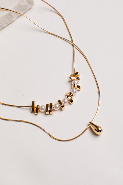 A gold-toned double-strand necklace featuring a central cluster of small white pearls and gold spacer beads, with a teardrop-shaped gold bead pendant, laid out on a light neutral background.
