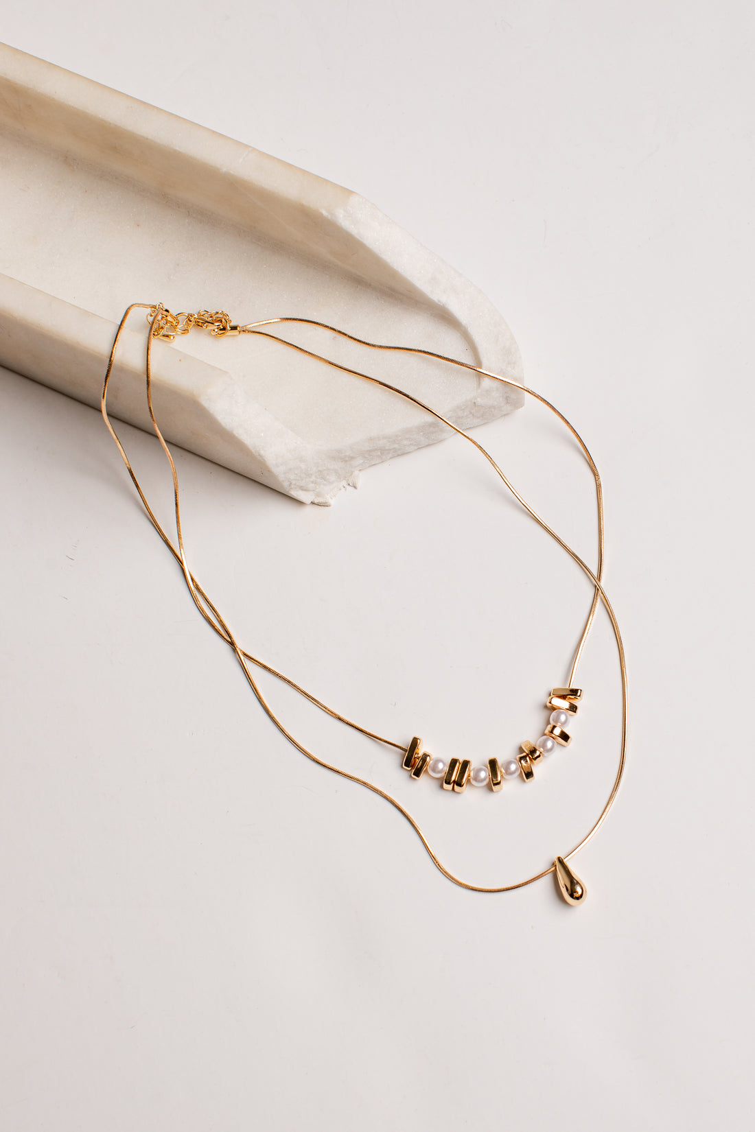 A delicate gold double-layer necklace with a small cluster of clear rhinestones and a tiny teardrop charm, displayed on a light beige textured ceramic prop against a white background.