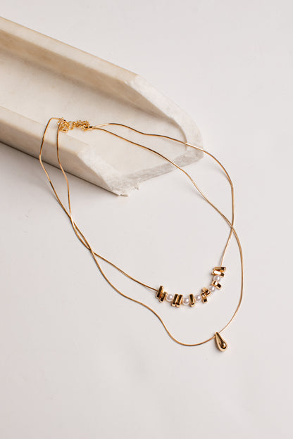 A delicate gold double-layer necklace with a small cluster of clear rhinestones and a tiny teardrop charm, displayed on a light beige textured ceramic prop against a white background.