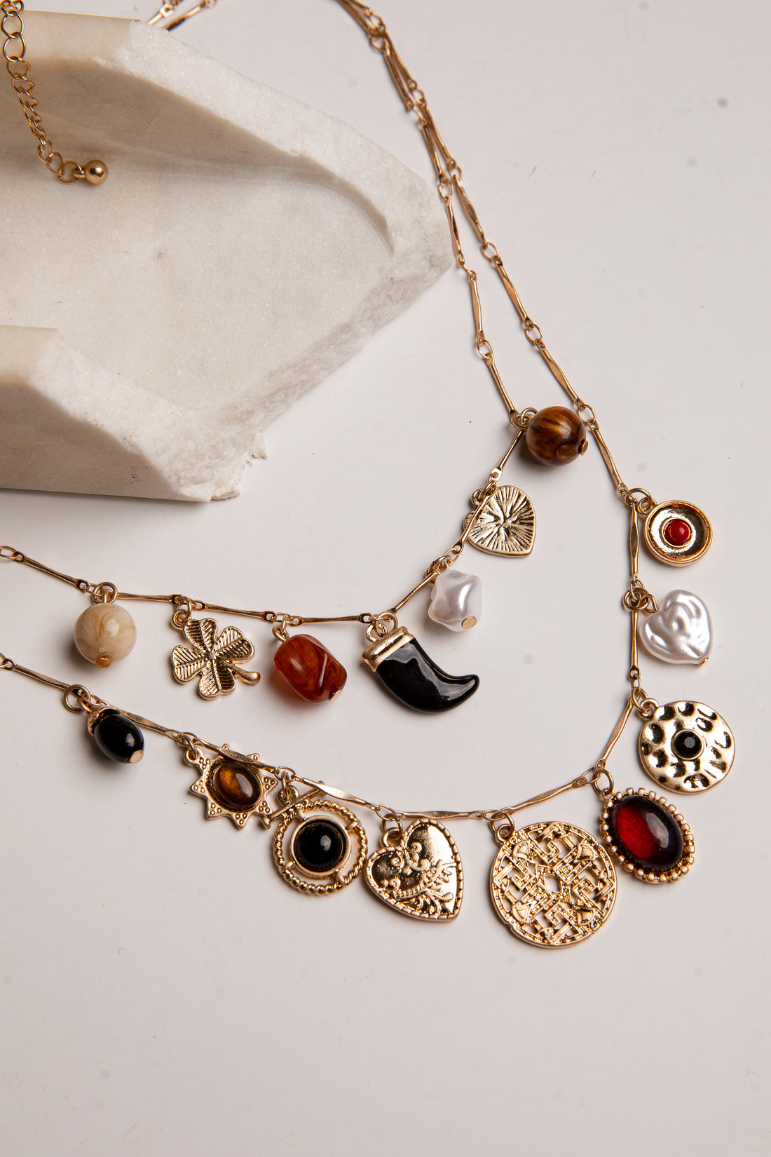 A gold-toned multi-strand necklace with various charms, including a clover, crescent moon, hearts, and round cabochon stones in red, black, brown, and white, displayed on a light beige surface next to a white stone prop.