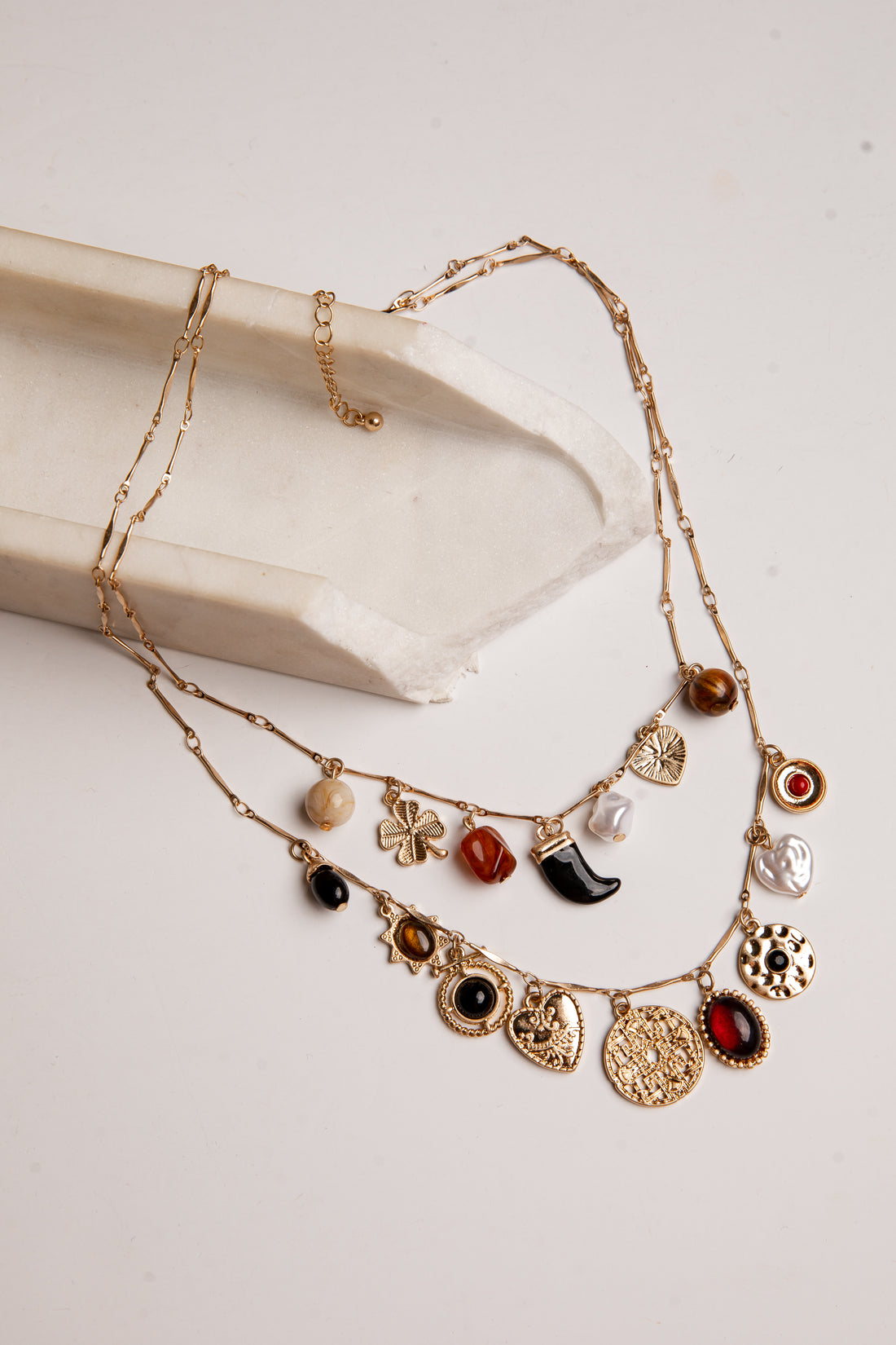 A gold-toned multi-strand necklace with various charms, including hearts, flowers, a crescent moon, and round beads, displayed on a cream-colored stone prop against a light beige background.