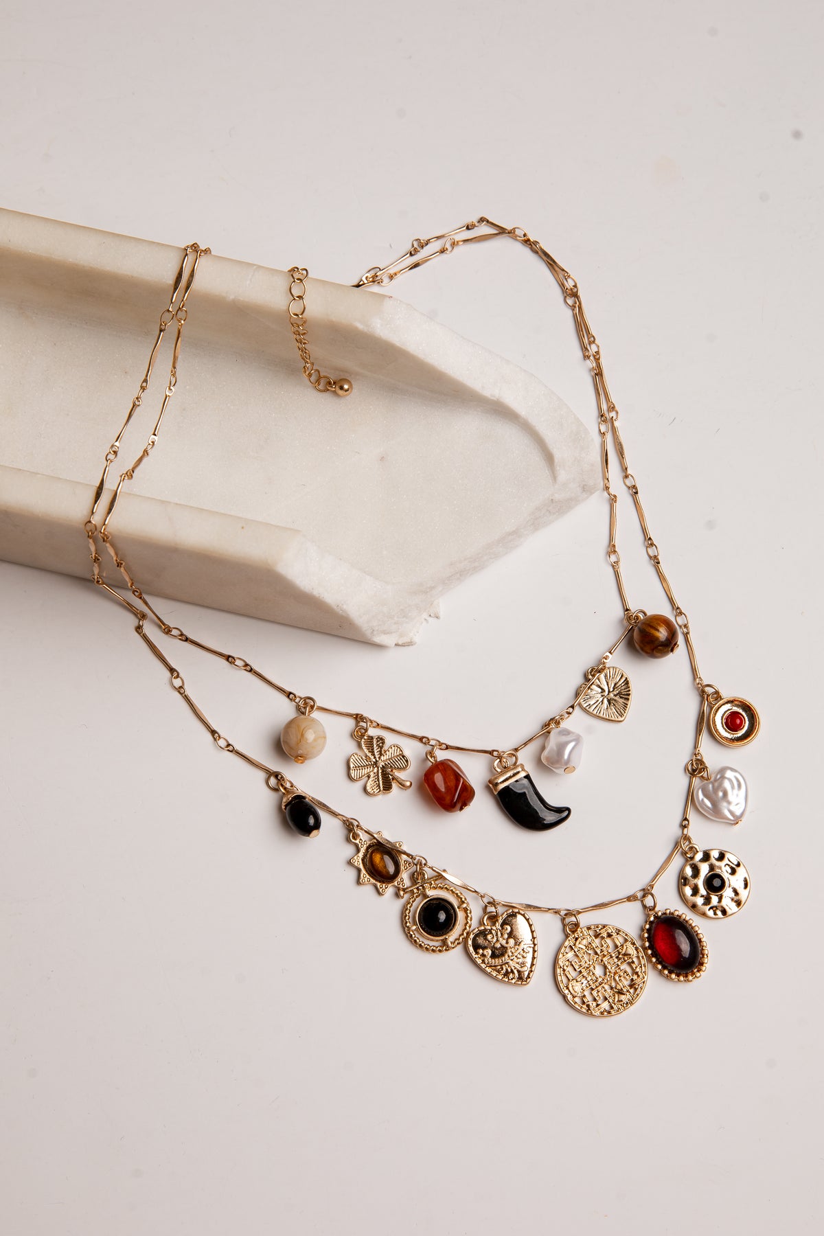 A gold-toned multi-strand necklace with various charms, including hearts, flowers, a crescent moon, and round beads, displayed on a cream-colored stone prop against a light beige background.