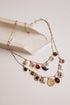 A gold-toned multi-strand necklace with various charms, including hearts, flowers, a crescent moon, and round beads, displayed on a cream-colored stone prop against a light beige background.