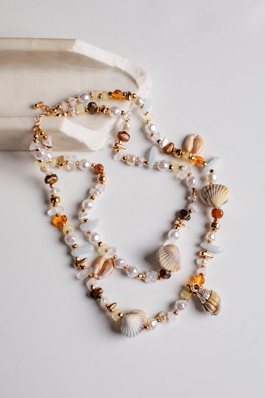 A multi-strand beaded necklace with seashells, white pearls, amber-colored beads, and gold-toned metal accents, styled on a light gray surface with a white stone prop.