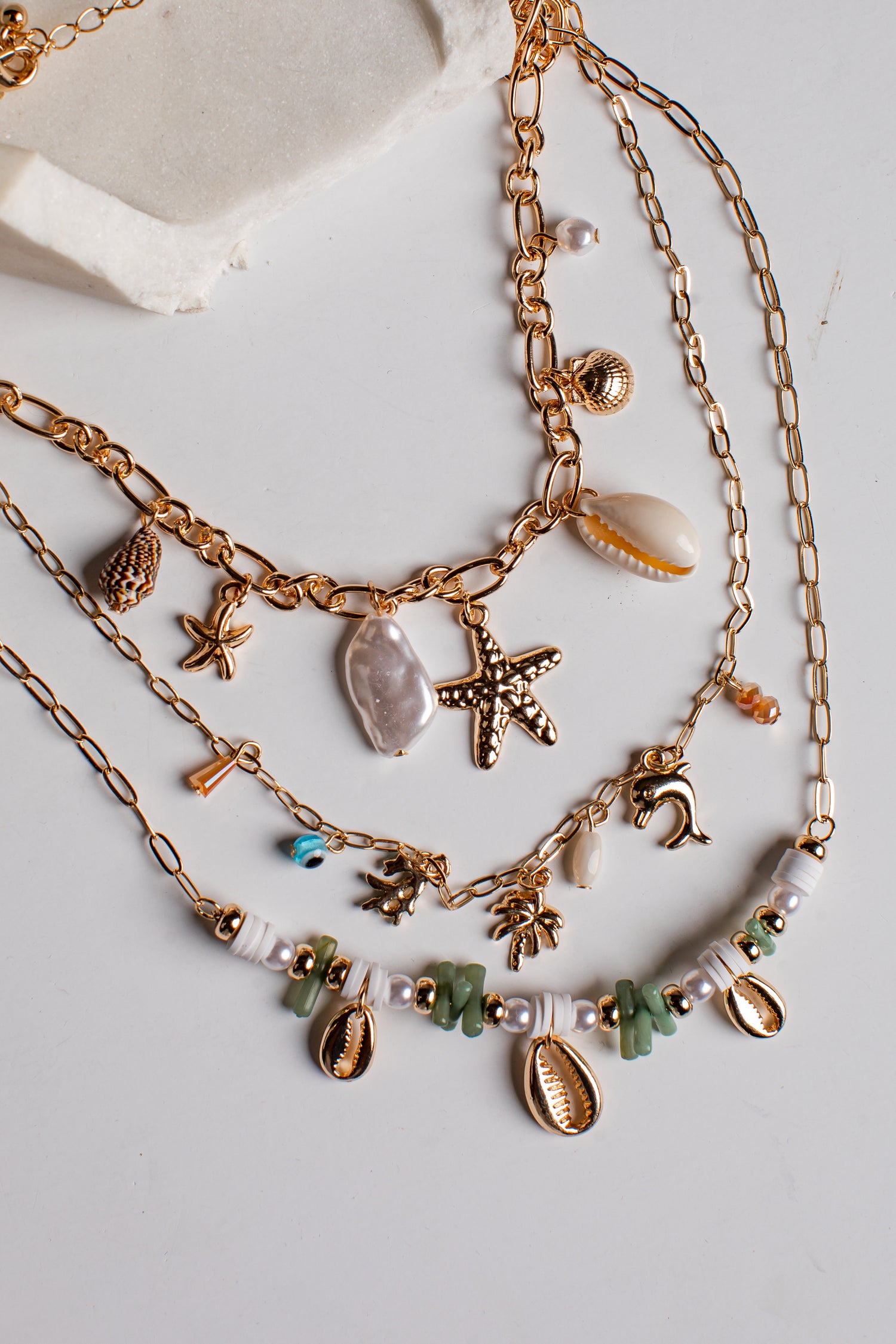 A layered gold-tone necklace set with seashell and starfish charms, displayed on a light gray surface with a textured white stone prop.