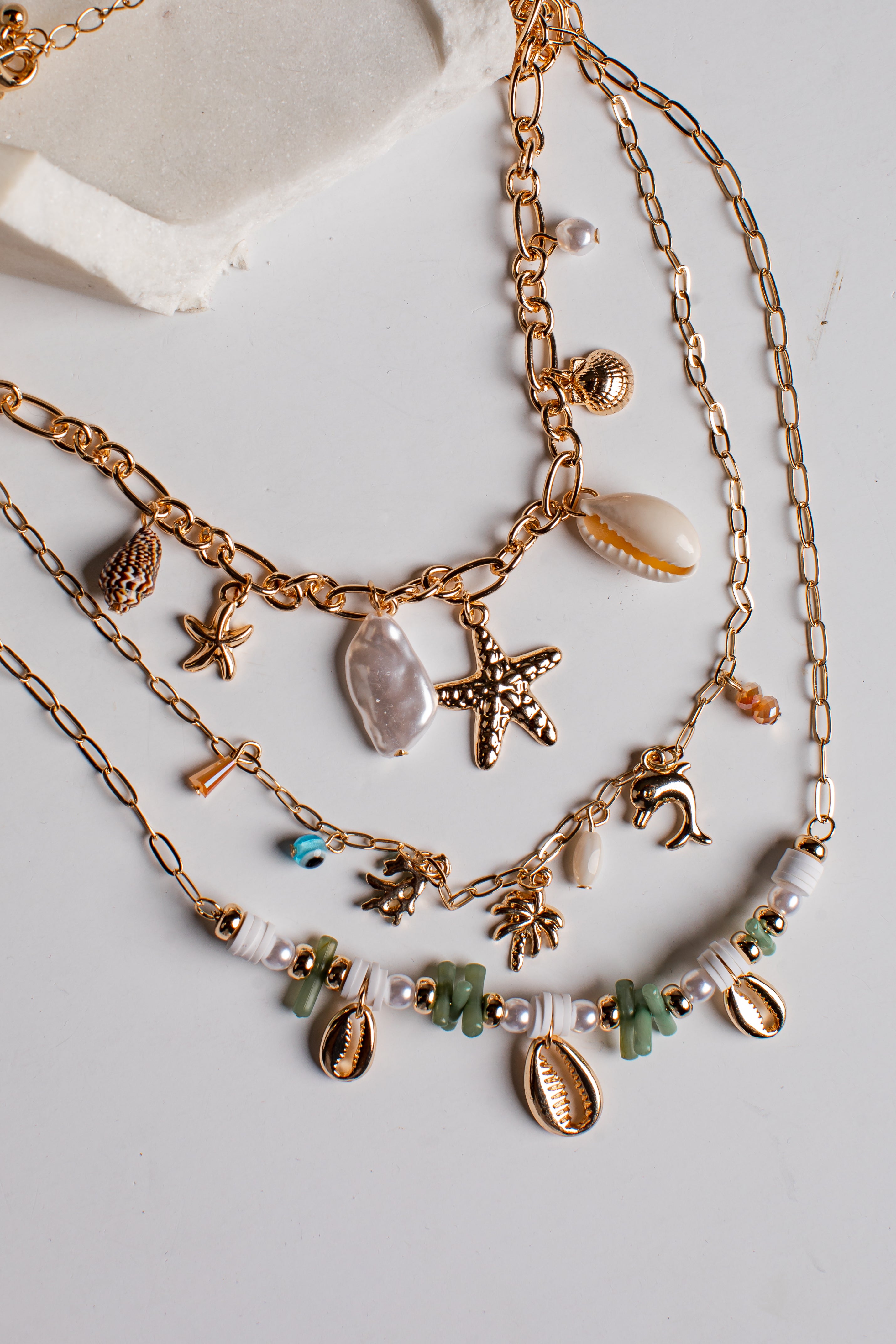 A layered gold-tone necklace set with seashell and starfish charms, displayed on a light gray surface with a textured white stone prop.