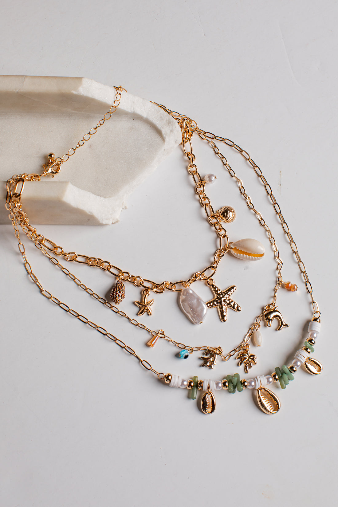 A multi-strand bohemian-style gold chain necklace with beach-themed charms including starfish, seashells, beads, and small sea creatures, displayed on a light gray surface next to a white stone prop.