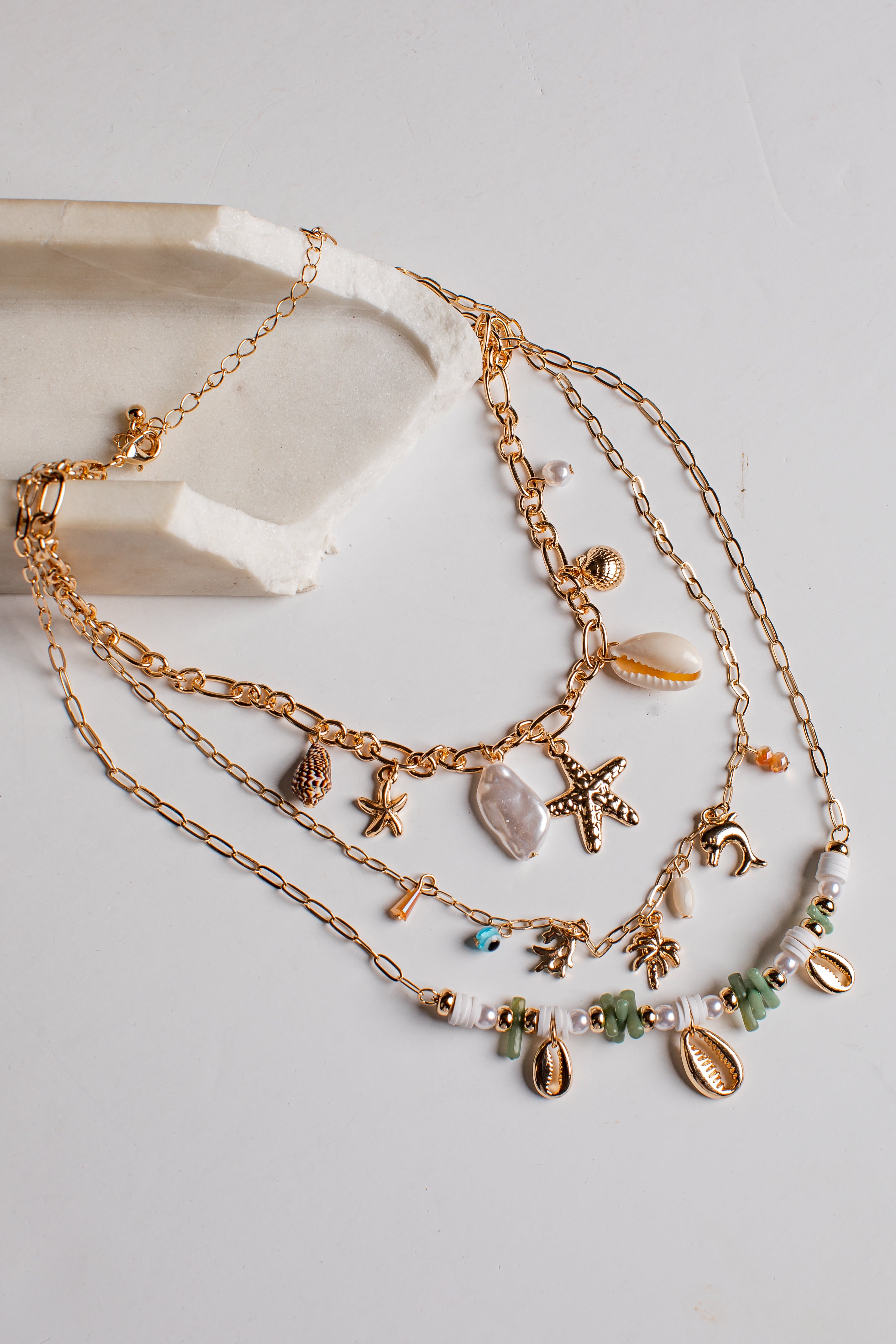 A multi-strand bohemian-style gold chain necklace with beach-themed charms including starfish, seashells, beads, and small sea creatures, displayed on a light gray surface next to a white stone prop.