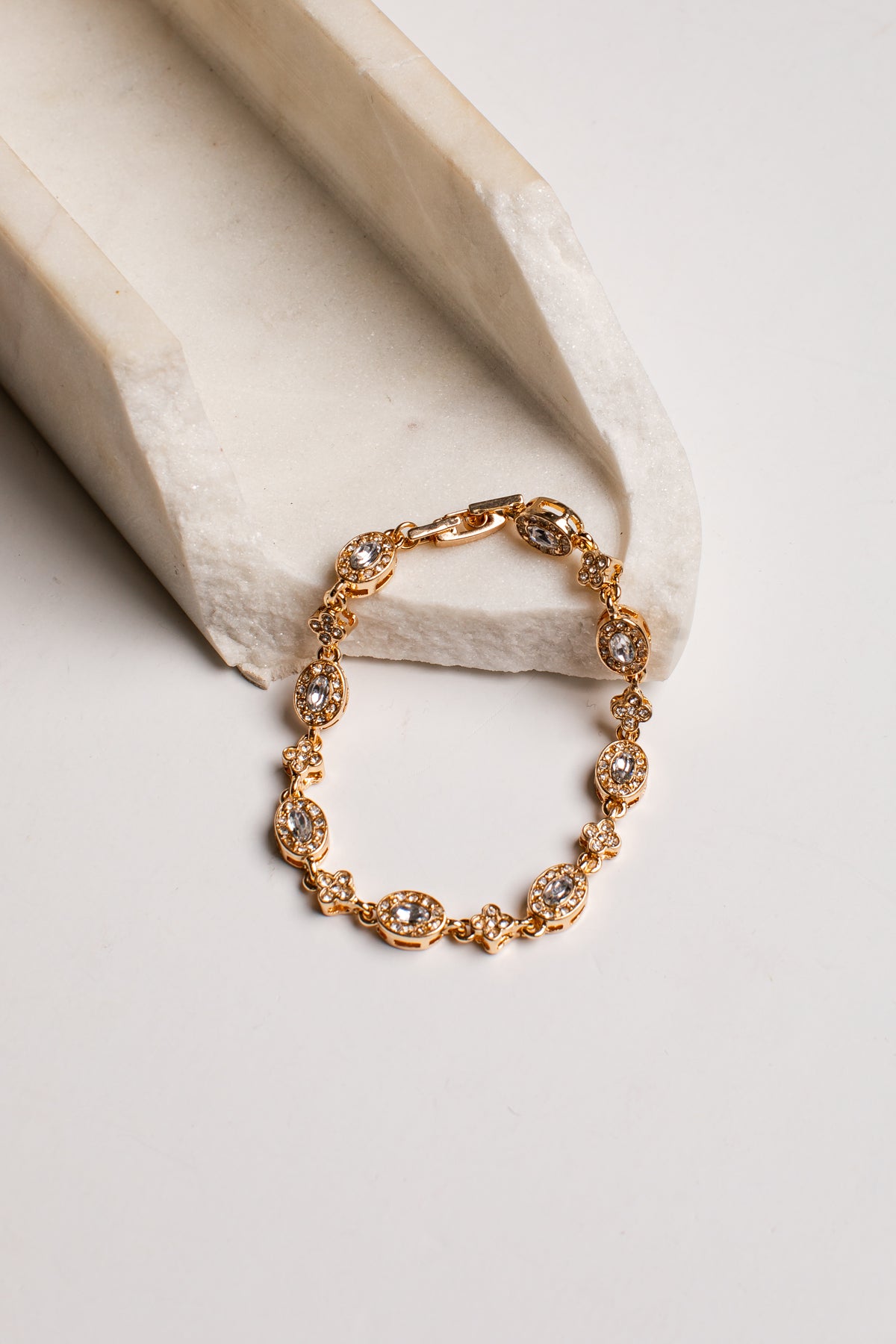 A gold-toned bracelet with multiple oval clear gemstones, displayed on a light cream stone riser against a clean off-white background.