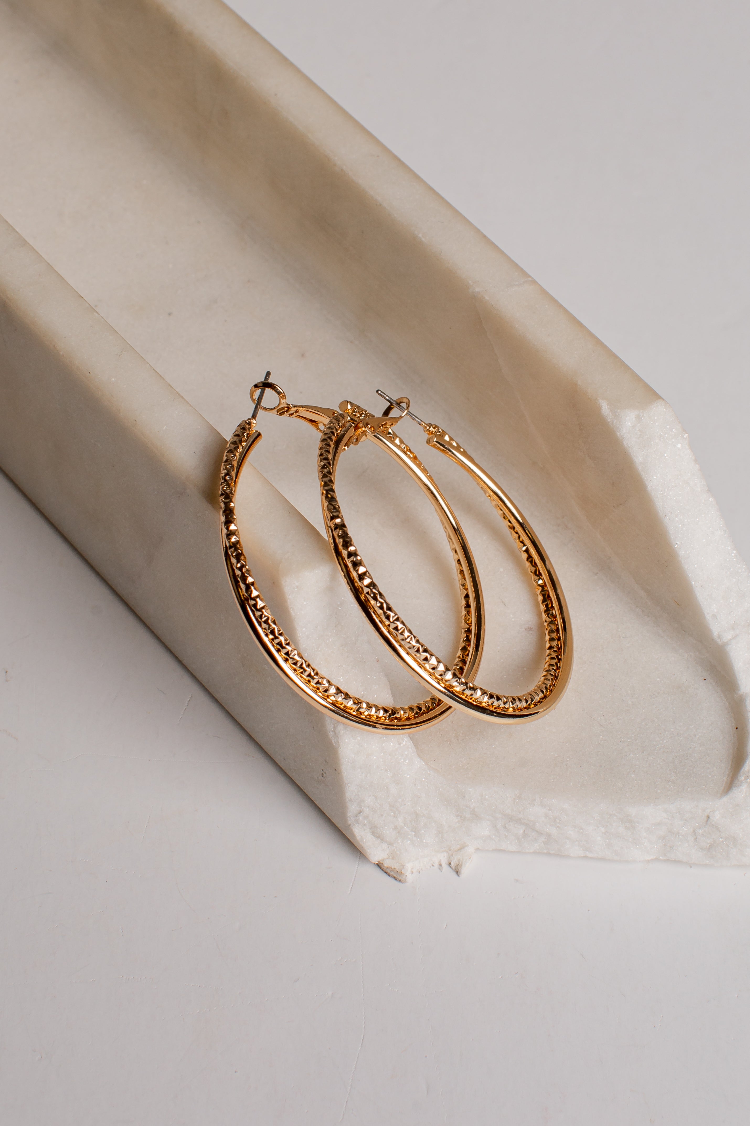 Gold hoop earrings with a textured, beaded outer surface and small fishhook backings, displayed on a white textured paper surface next to a cream-colored stone prop.