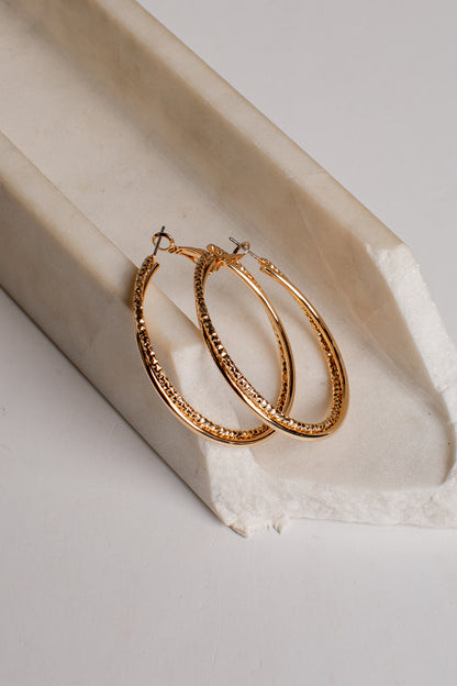 Gold hoop earrings with a textured, beaded outer surface and small fishhook backings, displayed on a white textured paper surface next to a cream-colored stone prop.