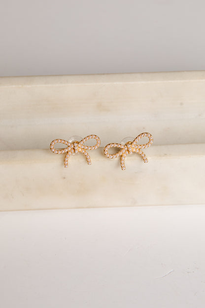Gold bow-shaped earrings on a beige cushion with a light gray background