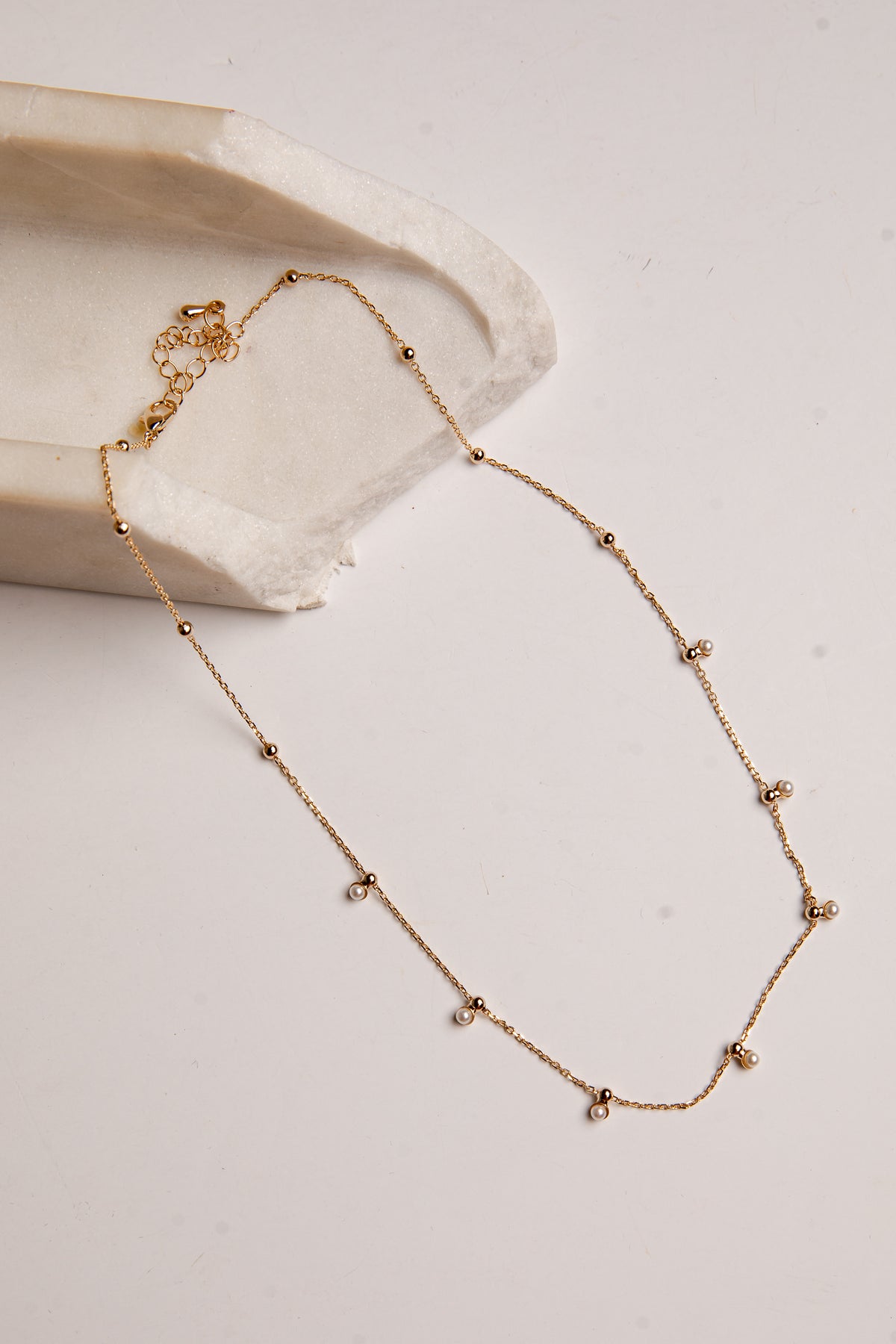 A delicate gold chain necklace with small bead accents and tiny clear stones, displayed on a light beige surface next to a textured cream stone prop.
